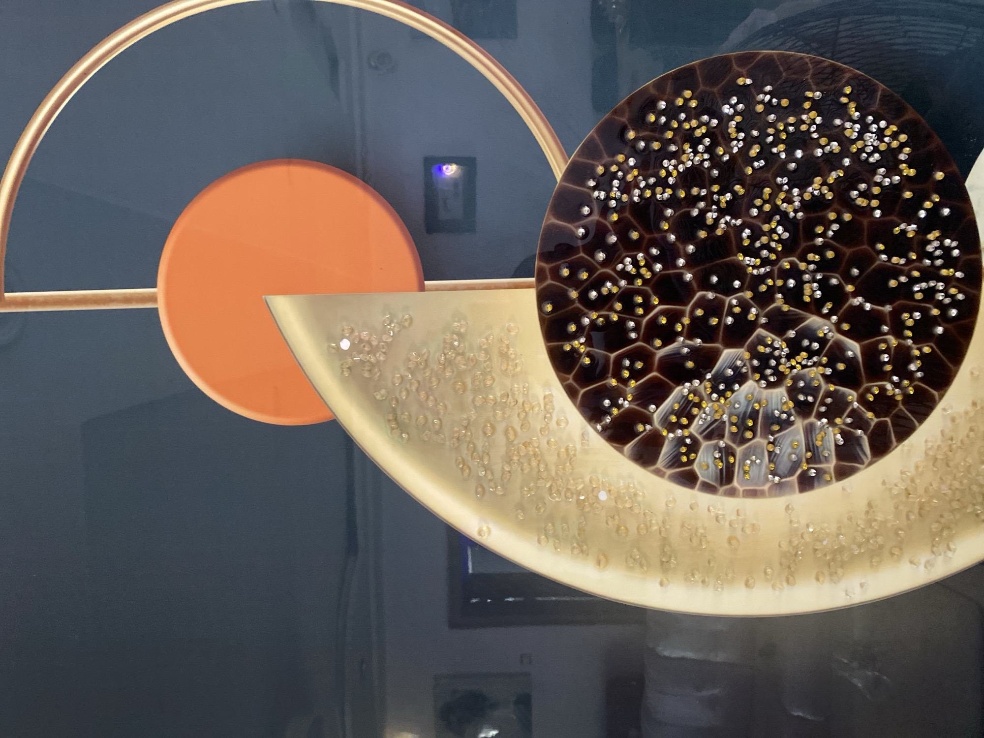 This image appears to be a close-up of a decorative art piece. The primary subjects are geometric shapes and textured surfaces. There is a large, dark brown circular object with a honeycomb-like pattern, speckled with numerous small gold and silver dots. To its left, a large, curved, metallic gold surface is visible, also adorned with many small, translucent, bead-like embellishments. Further to the left, a solid orange circle is positioned against a dark grey background, intersected by a thin gold arc.
No people are visible in the image. The activity taking place is not evident, as it is a still image of an object. The setting is ambiguous, but the abstract nature of the artwork suggests it could be displayed in an interior space, such as a gallery, home, or commercial establishment. There are reflections on the surfaces, indicating the presence of ambient lighting, but no specific time of day or weather conditions can be determined. No text is visible in the image.
