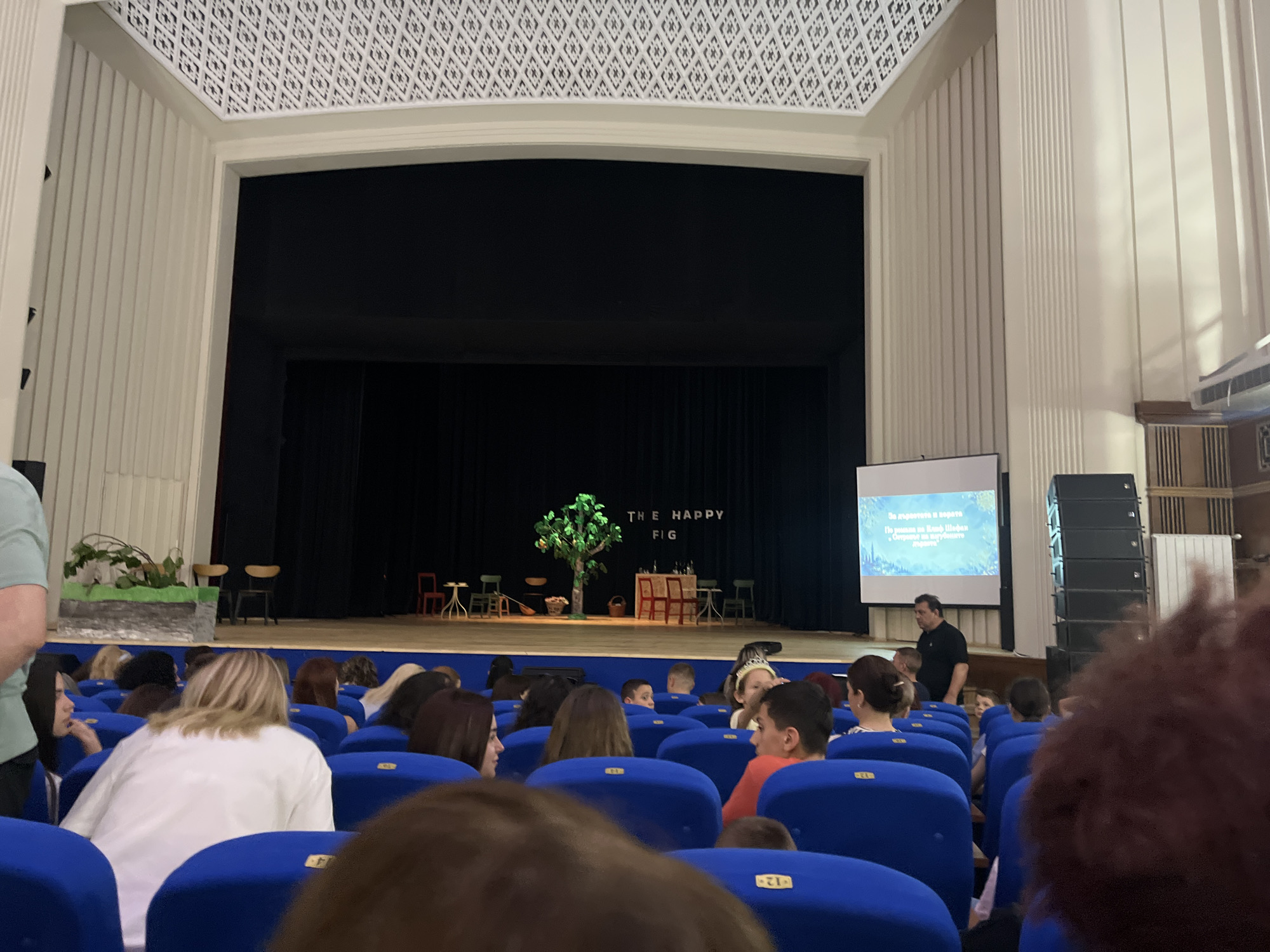An indoor photograph captures the audience and stage of a theater or auditorium in Asenovgrad, Bulgaria. The perspective is from the back of the audience, looking forward towards the stage.
In the foreground and midground, numerous people, including both adults and children, are seated in rows of blue upholstered chairs. Most audience members have their backs or sides to the camera, indicating they are facing the stage. One young boy in an orange shirt and a girl wearing a small crown are visible among the attendees.
The stage features a simple set with a black curtain backdrop. Props visible on stage include an artificial tree with green leaves, a small table with two chairs, another table with a red chair and a green chair, a broom, a basket, and a planter with green foliage. The words "THE HAPPY FIG" are displayed in white letters on the black backdrop.
To the right of the stage, a large projector screen displays text in Bulgarian. The text reads: "За дърветата и хората" (For trees and people), "По романа на Клиф Шефан" (Based on the novel by Cliff Sheffan), and "Островът на изгубените" (The Island of the Lost), suggesting a presentation or performance related to a book. A man in a black shirt stands near the screen, and a large stack of black speakers is positioned beside it.
The venue's architecture includes light-colored paneled walls and a distinctive white ceiling with a patterned black geometric design. The scene depicts an event, likely a play or lecture, in progress or about to begin, with attendees gathered in the performance hall.