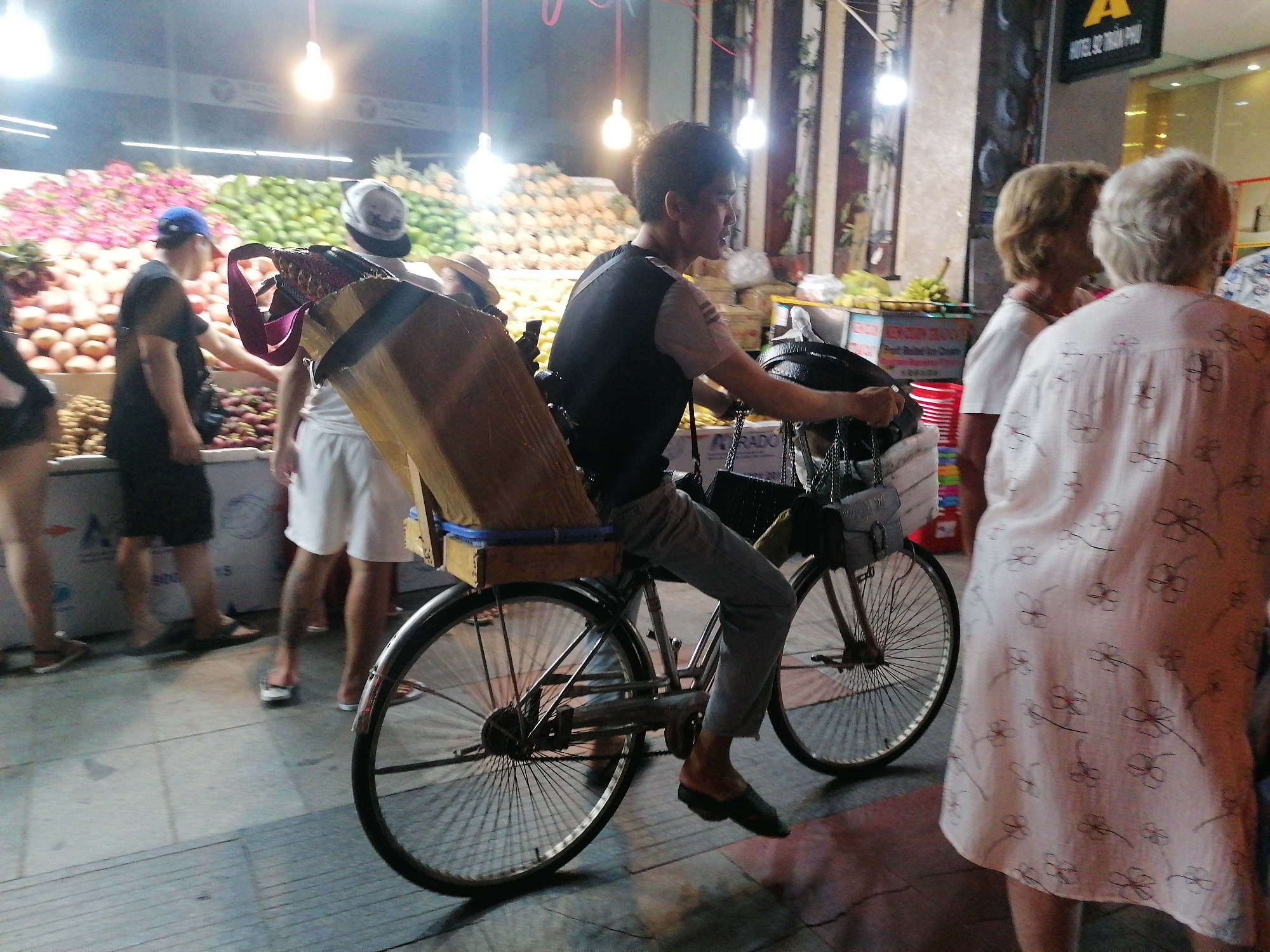 The image depicts a street scene in Nha Trang Ward, Vietnam. A man is riding a bicycle, which appears to be heavily laden with goods, past a fruit stand. The fruit stand displays a variety of fruits, including what appears to be mangoes and dragon fruit. Several people are standing near the fruit stand, seemingly browsing the produce. In the foreground, two women are partially visible, facing away from the camera, observing the fruit stand. The scene is lit by overhead lights.