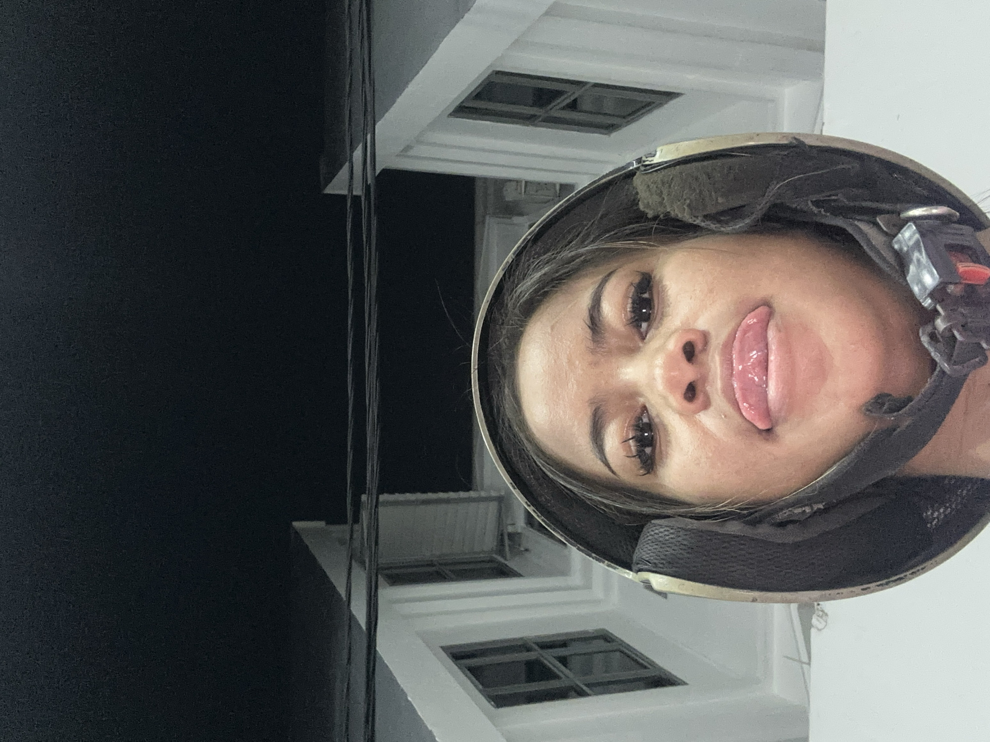 A young woman is wearing a helmet and sticking out her tongue. The background shows the exterior of a building with windows. The location is likely Central Jakarta, Indonesia, based on available information.