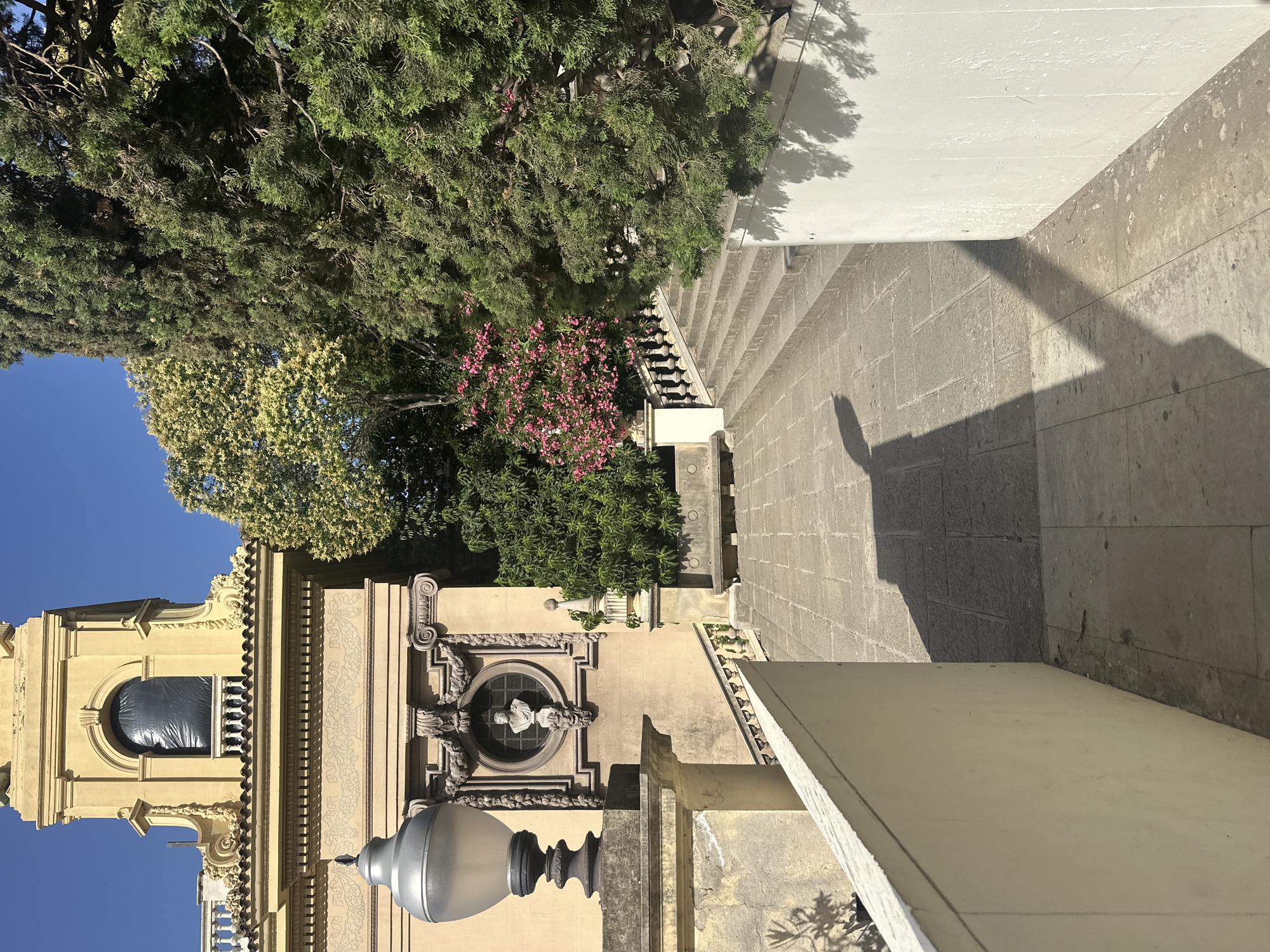 The image depicts an exterior view of a building with a prominent yellow facade, characteristic of Spanish architecture. The building appears to be a historical or religious structure, possibly a church or a bell tower, with ornate architectural details such as columns, carvings, and a rounded arch window. The window itself is covered with black plastic. A sculpture of a bust is visible within a circular niche on the facade.
In the foreground, a large, modern-style streetlamp with a silver-toned shade stands out. The ground is paved with large stone tiles, and a set of stone steps leads up to a higher level. Lush green trees and a flowering bush with pink blossoms are visible on the left side of the image, providing a natural contrast to the built environment.
The scene is bathed in bright sunlight, casting strong shadows across the paving stones and the white wall of an adjacent structure. This suggests it is a sunny day, likely during the daytime. The clear blue sky further reinforces the impression of fair weather. The overall impression is one of a tranquil, sun-drenched environment, possibly a public square or a courtyard adjacent to a significant building in Barcelona, Spain. There is no visible text in the image.