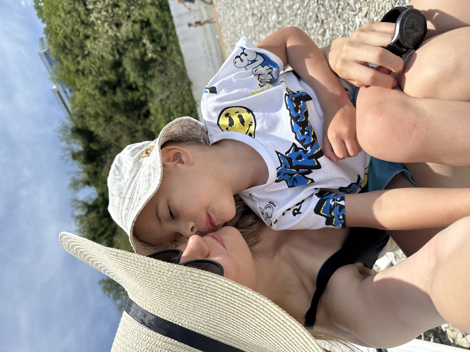 A blonde woman, partially visible on the left, wearing a large beige straw sunhat with a black band and dark sunglasses, leans in to kiss a young boy. She is wearing a black swimsuit or top. The boy, positioned centrally, wears a light-colored bucket hat with a small brown emblem and a white t-shirt adorned with yellow smiley faces and blue graffiti-style text. He has fair skin and is holding a black smartwatch in his left hand, which rests on his knee. Both subjects are on a pebble beach under a partly cloudy blue sky in Nebug, Russia. In the mid-ground, a low, white wall runs horizontally, with two blurred figures walking on the beach beyond it. The background features a steep, densely tree-covered hillside, with a modern multi-story building featuring large windows visible at the top of the hill.