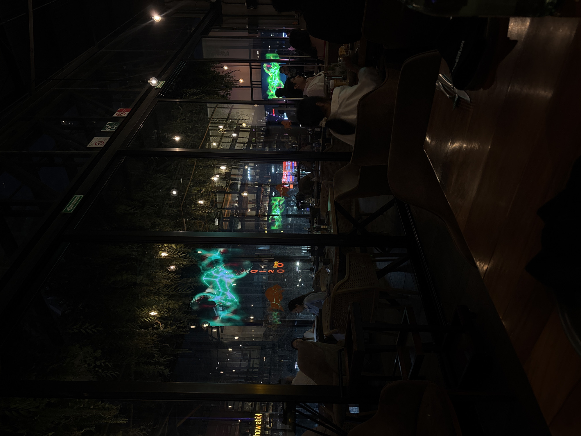 The image depicts the interior of a cafe or restaurant, seemingly at night, with a partial view of a city through a large window. Several people are visible, some seated at tables. The exterior scene shows city lights and possibly a television screen displaying an image in shades of green. The interior is dimly lit, with a focus on the tables and chairs. It has been confirmed that this photo was taken in Puebla, Mexico.