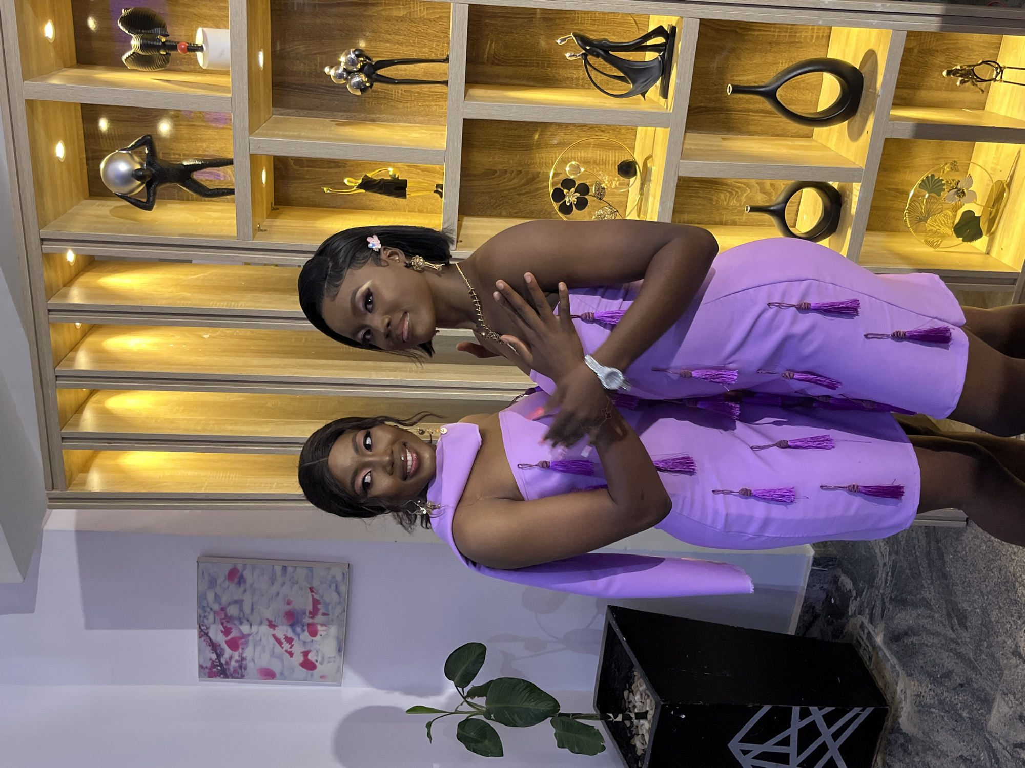 Two young Black women, both wearing matching lavender off-the-shoulder dresses with purple tassels, pose for a photo in an indoor setting. The woman on the left is looking to her right and smiling, with her right arm extended forward, while the woman on the right is looking towards the camera with her hands crossed over her chest. The dresses appear to be made of a smooth fabric, possibly satin, and are sleeveless.
They are standing in front of a large wooden shelving unit filled with various decorative items. These include abstract sculptures, vases, and what appear to be figurines. The shelves are illuminated by warm, recessed lighting, creating a bright and airy ambiance. To the left of the women, there is a large black planter containing a leafy green plant. The wall behind the plant is a light, neutral color, and a framed piece of art depicting pink koi fish is hanging on it. The floor appears to be made of polished stone with a mottled grey and black pattern. The overall scene suggests a modern, well-decorated interior, possibly a home or a boutique. The individuals and decor are well-lit, making it easy to discern details. The location is Gura Top, Nigeria.