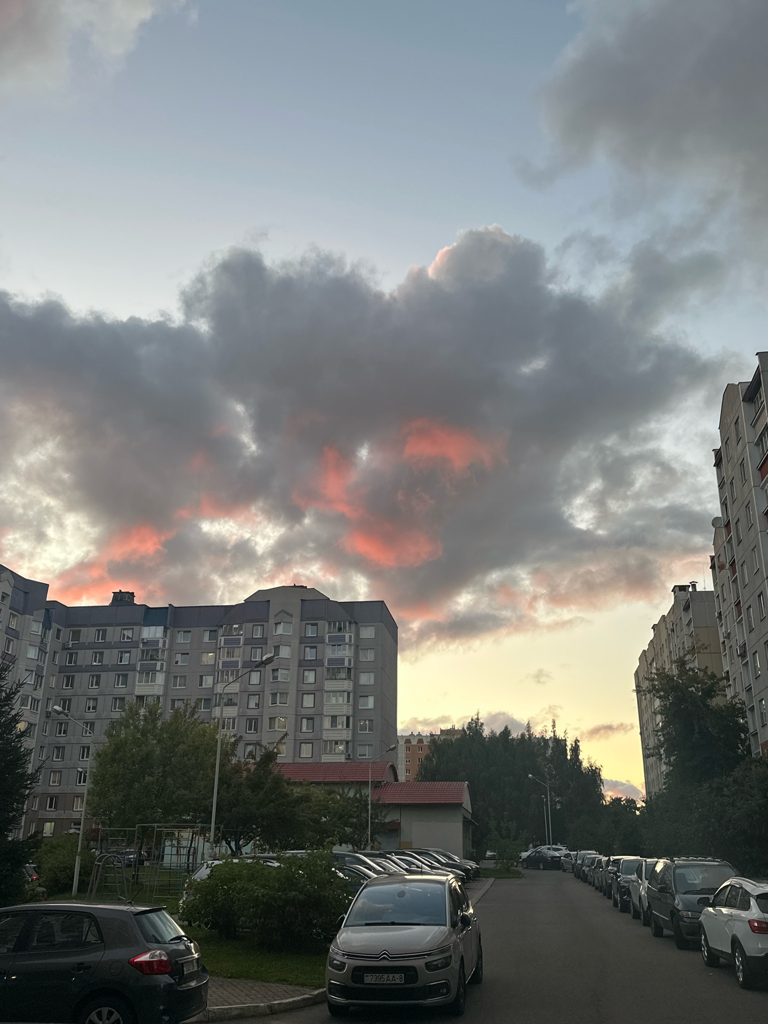 This is a vertical, eye-level, outdoor shot taken in a residential courtyard in Minsk, Belarus, during what appears to be sunset or dusk. The sky is filled with dramatic, gray clouds tinged with pink and orange hues, suggesting the sun is low on the horizon. In the middle ground, multiple large, multi-story apartment buildings, typical of Soviet-era architecture, dominate the scene. These buildings are made of gray concrete and have numerous windows. Between the buildings, trees and some low-rise structures, including a building with a red-tiled roof, are visible. The foreground shows a paved courtyard with a row of parked cars, mostly sedans and SUVs. A lamppost is also visible in the courtyard. The overall impression is of a typical urban residential area experiencing a colorful sky.