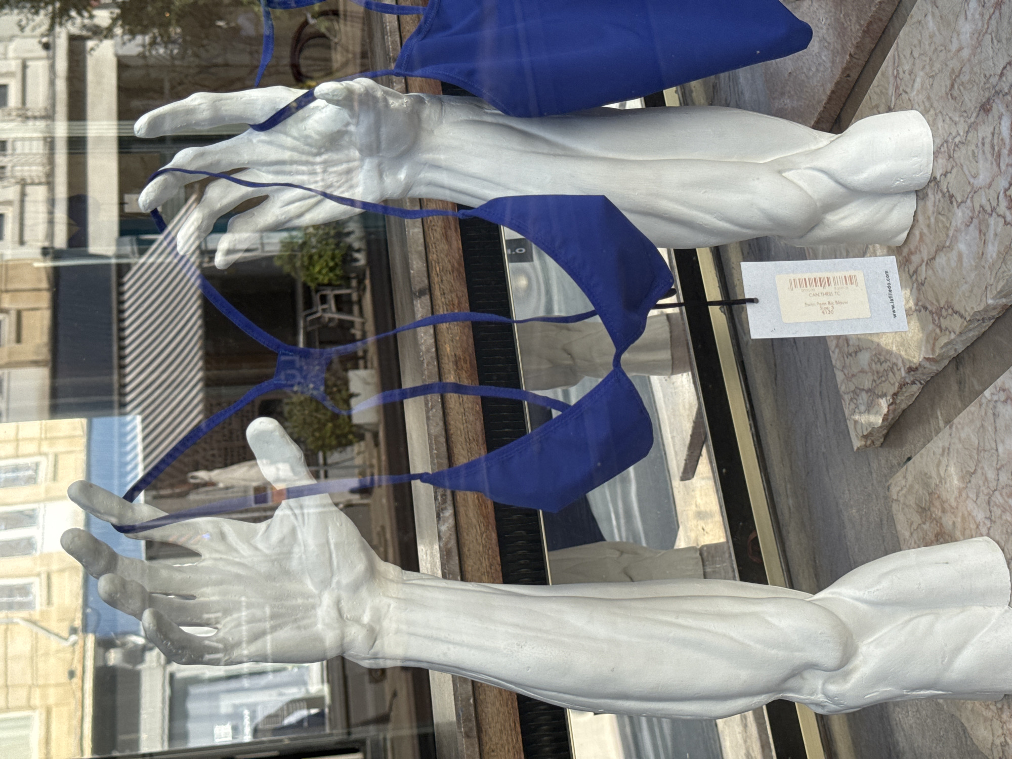 This image shows a display in a shop window on a sunny day. Two white plaster arm and hand mannequins are on display, each holding a blue bikini top. The mannequins are positioned in front of a shop window that reflects the street outside, showing buildings with windows and awnings. A price tag is attached to the left mannequin. The display is set on marble slabs. The reflection in the window includes a striped awning and a building with multiple windows. The overall scene appears to be a retail display for swimwear. The city of Antwerp, Belgium is mentioned in the context of this image.