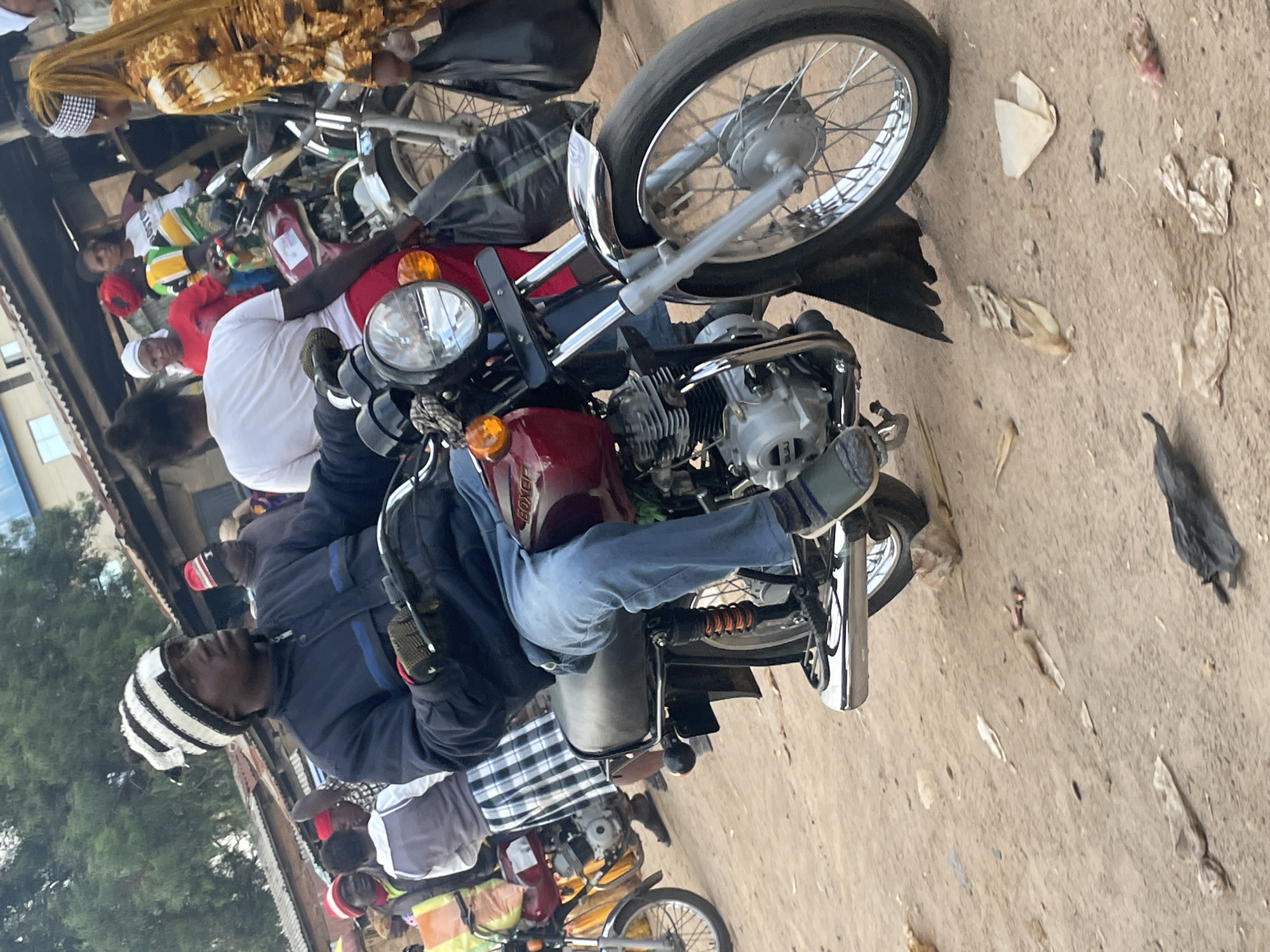 A man wearing a black and white striped beanie, a dark blue jacket, and blue jeans rides a red motorcycle in a dusty, outdoor market. He is wearing green shoes. Several people are gathered around him, some on motorcycles, in the background. The scene appears to be in Wang, Nigeria.