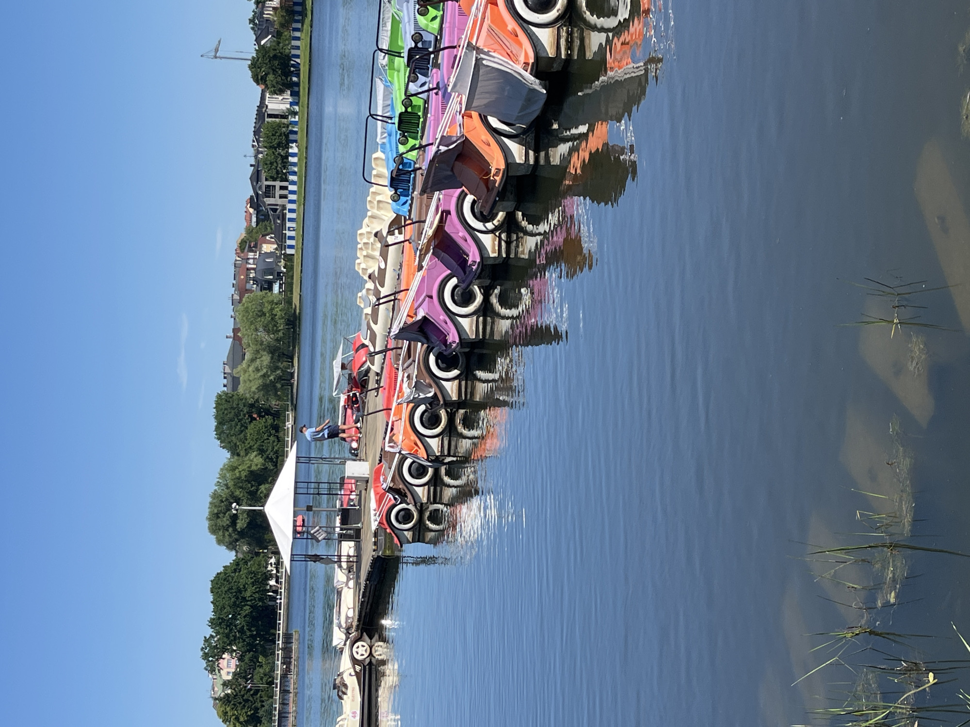 The image shows a row of colorful pedal boats docked on a clear blue lake on a sunny day. The boats are various colors, including pink, orange, green, and blue. They are lined up along a wooden pier, with some people visible on the pier. One person in a blue shirt and shorts is standing near the edge of the pier, looking out at the water. The water reflects the boats and the surrounding buildings, which appear to be houses and businesses. The sky is clear and blue, indicating good weather. There is a crane visible in the distance, suggesting ongoing construction or development in the area. The scene appears to be a recreational area, likely a park or a waterfront attraction. The overall mood is peaceful and relaxed, fitting for a summer day. The image was likely taken in Kaliningrad, Russia, given the provided location context. No visible text is present in the image.