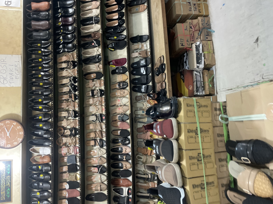 An interior view of a densely packed footwear retail establishment in Nganguk, Indonesia, is depicted. Multiple tiers of metal grid shelves and a wooden countertop display thousands of shoes. The upper shelves feature various styles of women's dress shoes, flats, heels, and sandals in colors predominantly black, brown, beige, white, and metallics. The lower displays showcase sneakers (including "BELBE" branded maroon, black, and beige pairs), chunky black loafers, and sandals/slides (some branded Nike and Adidas). A white sign with red lettering "GROSIR TIDAK ECER!" (Wholesale, not retail!) is affixed to the upper right wall. A brown wall clock displaying approximately 10:10 and a framed piece of green Islamic calligraphy are visible on the upper left wall. Numerous cardboard boxes, some bearing the "DesFiles" brand and handwritten labels like "N17" and "3cm," are stacked beneath the counter and on the light-colored concrete floor. No individuals are present in the scene.