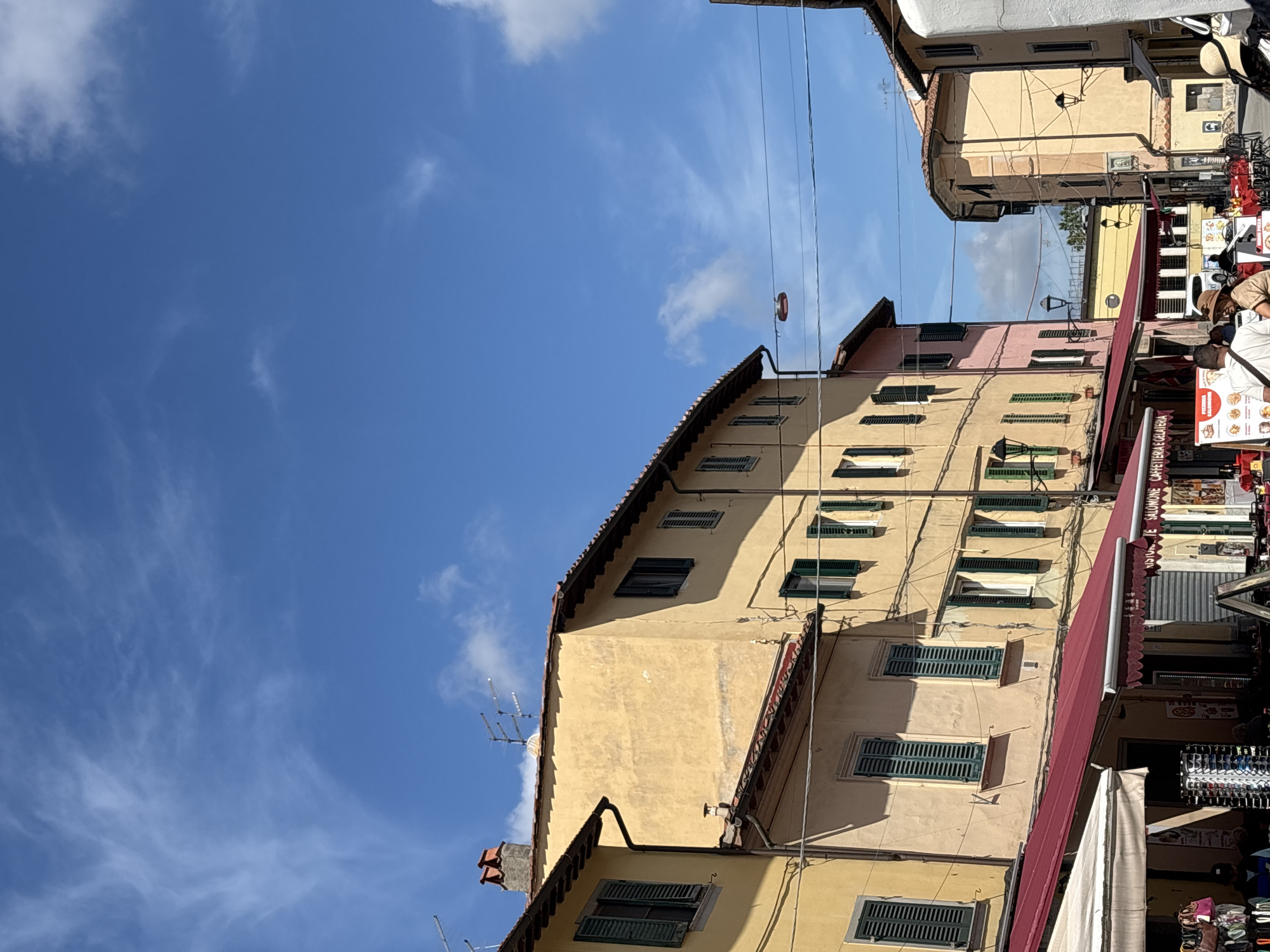 The image depicts a street scene in Pisa, Italy. Buildings with yellow and pink facades are visible, featuring green shutters on their windows. A red awning extends over what appears to be a market area with various stalls and vendors, including people, presumably tourists. The sky above is blue with scattered clouds. Power lines run across the scene. The architecture suggests a historical European setting.