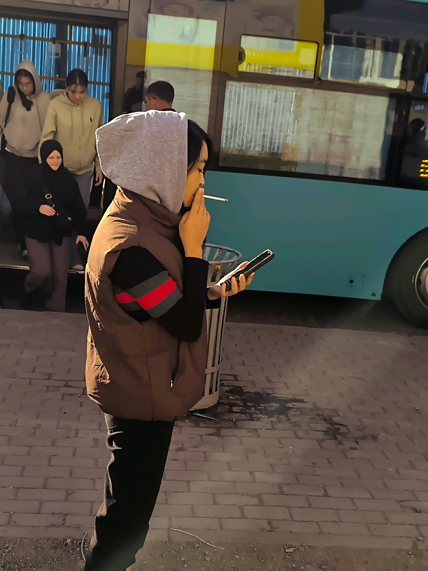In Qarağandı city administration, Kazakhstan, at street level, a young person wearing a brown puffer vest over a black long-sleeved shirt with red and green stripes on the sleeve, and a grey hood pulled up, stands near a bus. The person is holding a cigarette in their right hand near their mouth and a smartphone in their left hand. Behind them, other individuals are visible near what appears to be a bus entrance, with some ascending steps. A trash can is situated to the right of the main subject. The ground is paved with brick-like stones. A light blue bus is parked behind the individuals, with its side visible. The scene appears to be outdoors, possibly at a bus stop or terminal, during daylight hours with ambient lighting.