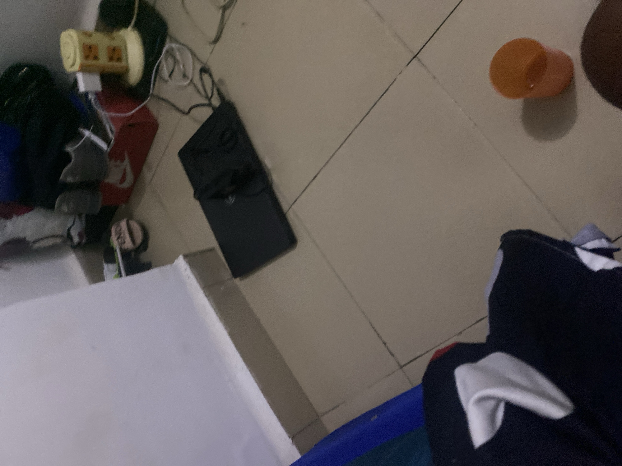 The image shows a cluttered floor in what appears to be a residential room, possibly in Howayi, Nigeria. In the foreground, a dark-colored fabric item with white detailing is partially visible. To the right, an orange cup casts a shadow on the tiled floor. In the mid-ground, a black laptop is open on the floor. Behind the laptop, a red Nike shoebox is visible, containing a pair of grey sneakers. A yellow cylindrical object, possibly a night light, is positioned to the right of the shoebox, connected to a power cord. Other items, including a pair of pink flip-flops and a black bag, are also scattered in the background, near a wall. The overall impression is one of disarray.