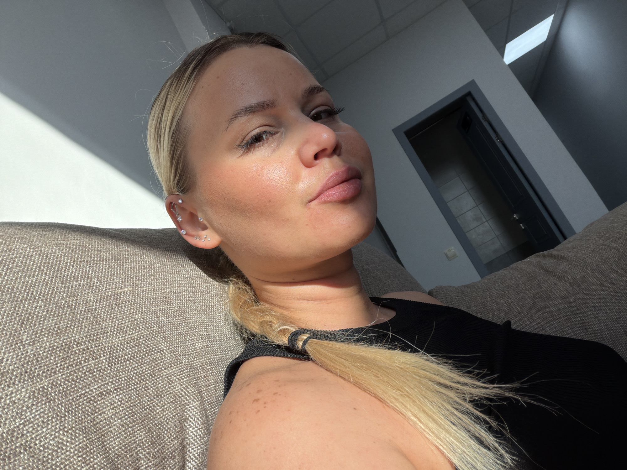 A medium close-up depicts a fair-skinned blonde woman with light eyebrows and light eyelashes, seated on a textured brown couch. She is wearing a black tank top and her hair is pulled back into a low ponytail, exposing multiple ear piercings. Sunlight illuminates her face from the left, creating strong shadows. The background includes a neutral-colored wall and a partially visible doorway. The specific location in Sarapul, Russia is not determinable from the visual content.