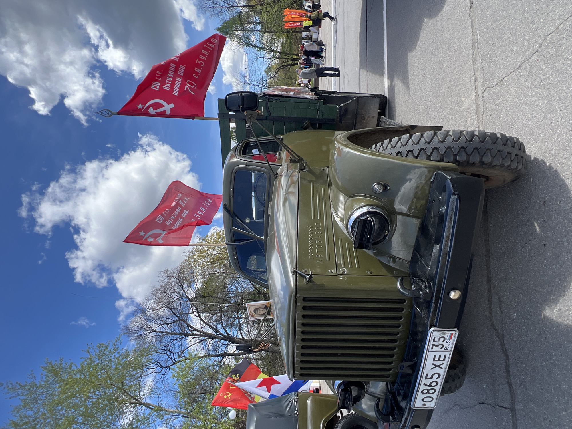 The image shows a vintage, olive-green truck with the license plate "O865 XE 35 RUS" parked on an asphalt road. The truck is adorned with red flags featuring the hammer and sickle symbol, and text in Russian. The flags also read "150 СТР. КУТУЗОВА Іст. ИДРИЦК. ДИВ." On the truck is also a sign that says, "ГОРЬКОВСКИ АВТОЗАВОД". In the background, there are more flags and a group of people standing on the side of the road. The sky is blue with white clouds.