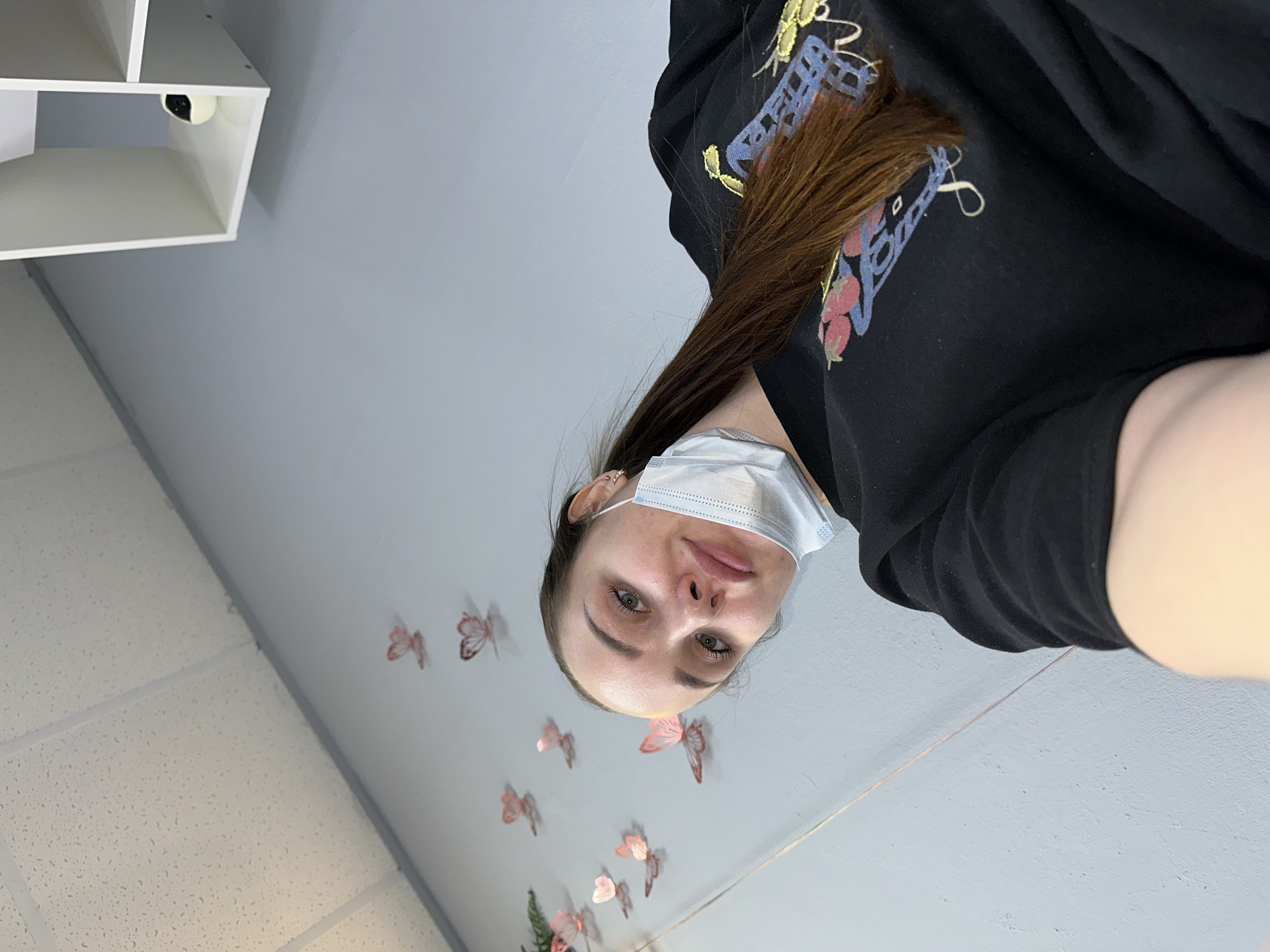 The image shows a person wearing a black hooded sweatshirt and a surgical mask, with brown hair. The person is looking into the camera. The background features a gray wall decorated with butterfly stickers and a glimpse of a white shelving unit in the upper left corner. The location is likely indoors, with fluorescent lighting. The image was likely taken in Chaikovsky, Russia.
