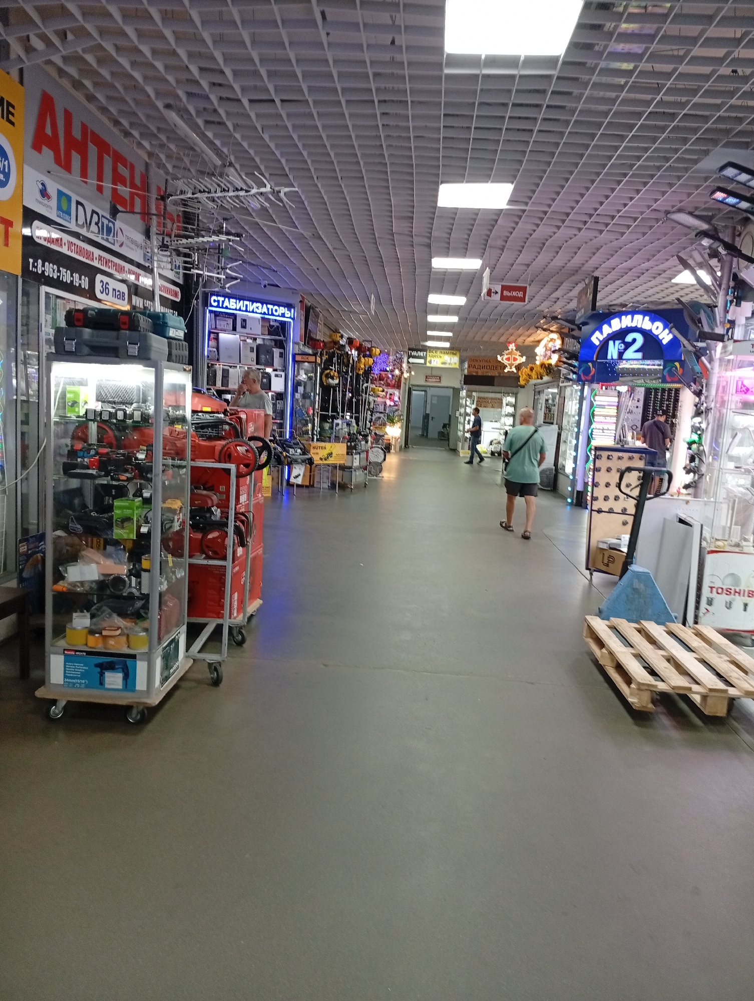 The image depicts the interior of a commercial establishment, likely a market or a large retail store, with an indoor setting. The ceiling is a grid of white tiles with fluorescent lighting. The floor is a plain gray surface.
In the foreground, to the left, is a glass display cabinet filled with tools and equipment, including power tools and toolboxes. To the right of the cabinet are several red wheeled carts loaded with more red power tools.
Further down the aisle, several people are visible. One person, wearing a green t-shirt and shorts, is walking away from the camera. Another person, wearing a dark t-shirt and pants, is standing and looking at products displayed in a brightly lit booth to the right, which has a sign reading "ПАВИЛЬОН №2" (Pavilion No. 2) in blue neon letters. Other individuals are present in the background, also browsing or working in the various stalls.
Various signs are visible throughout the scene. One large sign at the top left reads "АНТЕННЫ" (Antennas) in large red letters, along with other text in smaller font. Another sign further down the aisle says "СТАБИЛИЗАТОРЫ" (Stabilizers). A red sign with a white arrow points towards an exit, labeled "ВЫХОД" (Exit).
Other objects in the scene include pallets, shopping carts, and displays of merchandise. The overall impression is that of a busy marketplace selling tools and electronic equipment. The location is identified as Unknown, Unknown.