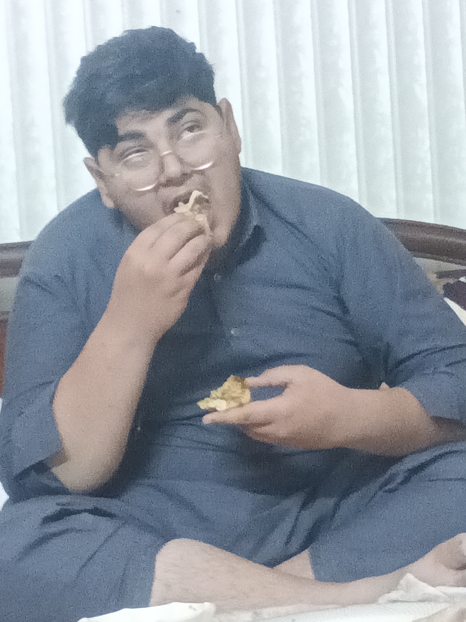 A male individual is depicted indoors, seated cross-legged, likely on a bed or cushioned surface. He appears to be of South Asian descent, with dark, curly hair and wearing eyeglasses. He is dressed in a loose-fitting, dark blue or gray traditional garment, consistent with a shalwar kameez.
The main activity captured is the man eating a snack. He holds a golden-brown, possibly fried, food item in his right hand, taking a bite, while another piece rests in his left hand. His mouth is open as he consumes the food.
The setting is an indoor environment, possibly a home in Samundri, Pakistan, as suggested by the cultural attire and provided context. The background features light-colored vertical blinds or curtains, indicating a window. A dark wooden headboard or furniture piece is partially visible on the right. The lighting is soft and even, suggesting either natural light from the window or ambient indoor lighting. The man's gaze is directed slightly upwards and to the right, possibly in thought or observing something off-camera while he eats. No visible text is present in the image.