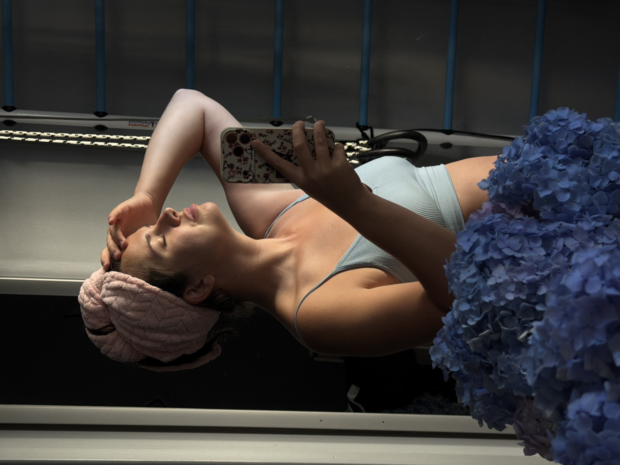 A young woman with a pink towel wrapped around her head and a light blue tank top is reflected in a mirror. She is holding a floral-patterned smartphone in her right hand, taking a selfie. Her eyes are closed and her hand is resting on her forehead. In the foreground, a large bouquet of blue hydrangeas is visible. Behind her, a wall with a ladder-like structure and blue bars can be seen. The lighting suggests it is daytime, with sunlight casting shadows. The scene is likely indoors, in a residential setting within Sochi, Russia.