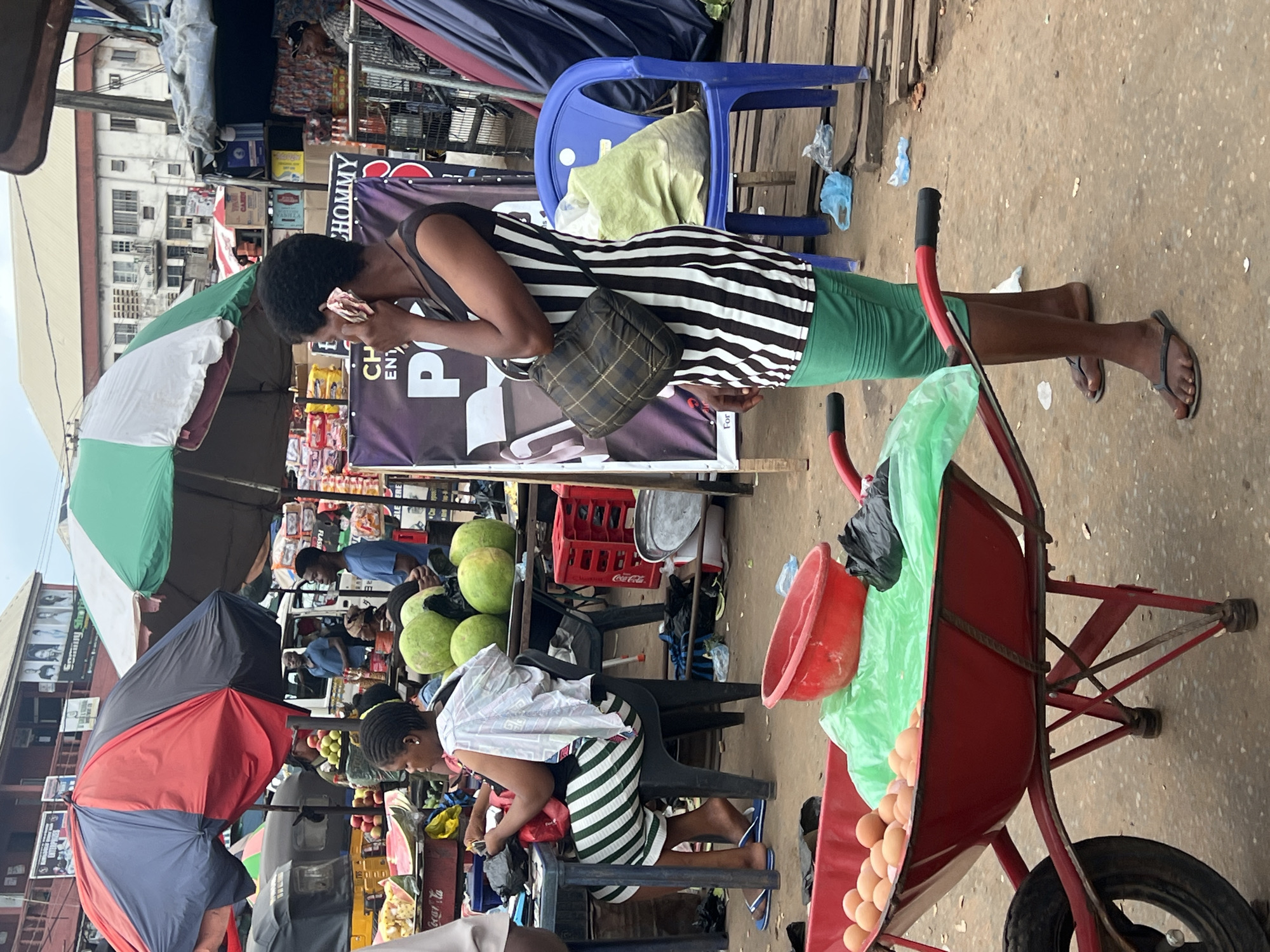 The image depicts a bustling outdoor market scene in Nkpor, Nigeria, likely during the day. A woman wearing a black and white striped top and a green skirt is seen in the foreground, holding a phone to her ear. She carries a checkered purse on her shoulder. Nearby, a red wheelbarrow is filled with eggs and a red basin. Behind her, another woman with braided hair is seated, wearing a striped skirt and top.
In the background, various market stalls are visible, adorned with colorful umbrellas. Stacks of green watermelons are arranged for sale, alongside other produce and goods displayed in crates. Several people are browsing or working at the market. A blue plastic chair is also visible, and the ground is a dusty, concrete surface. The overall impression is one of lively commerce. Some text is partially visible on signage behind the woman in the foreground, including "CHOMMY" and "PO". The weather appears to be fair.