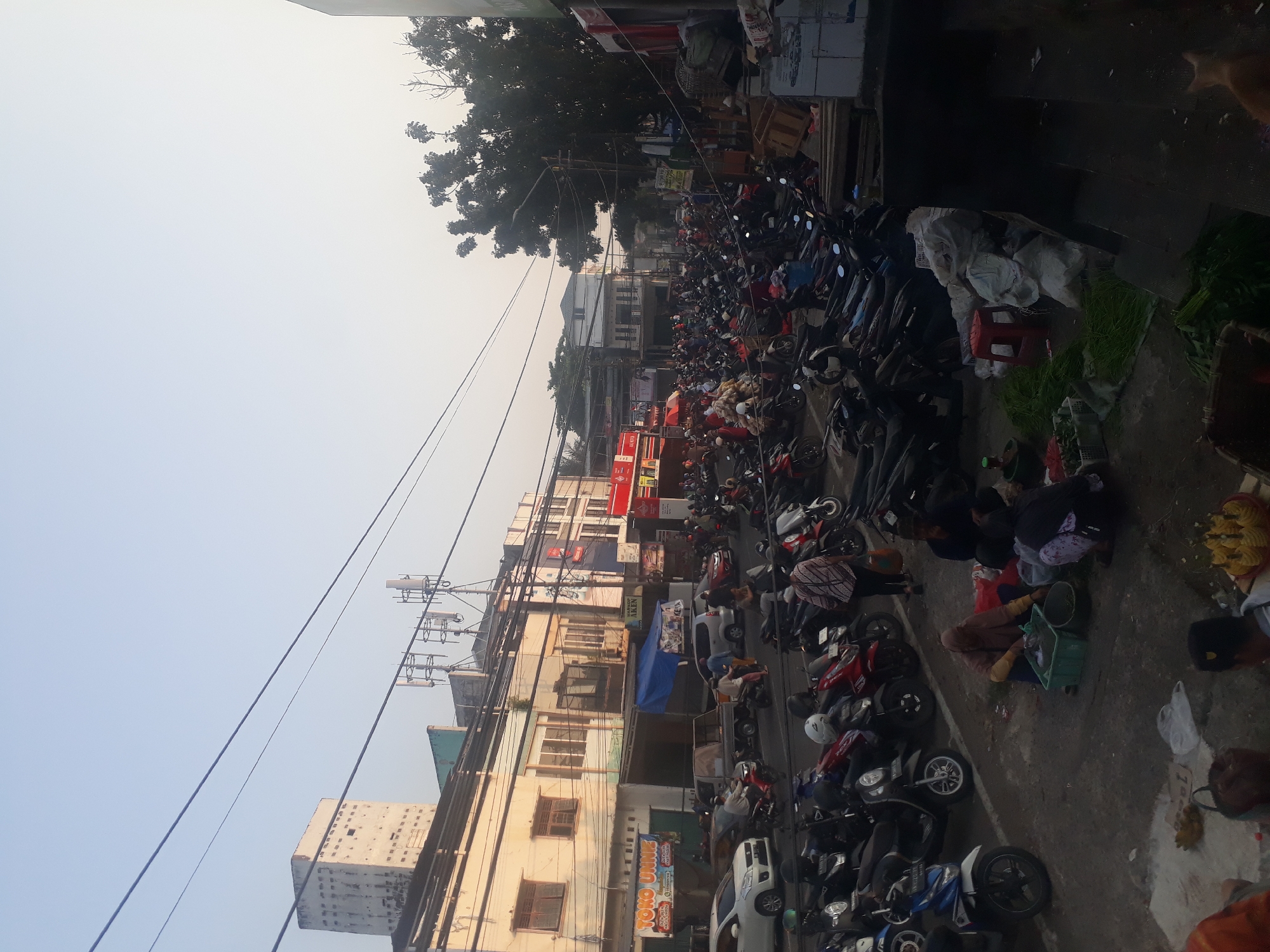 The image depicts a bustling street scene in a city, likely Pangkalpinang, Indonesia, characterized by a large number of parked motorcycles and several vendors. The time of day appears to be early morning or late afternoon, given the soft lighting and long shadows.
In the foreground, several individuals are engaged in selling goods. A woman in a hijab is tending to a green plastic crate filled with what appears to be fish or seafood, with another person kneeling beside her. Further to the right, another person is sitting on the ground, and a basket of produce, possibly pineapples, is visible. Scattered around are plastic bags, empty crates, and bundles of green leafy vegetables.
The middle ground is dominated by a dense parking area filled with motorcycles of various colors, suggesting a busy marketplace or transportation hub. Some motorcycles are occupied by riders, while others are parked. Several cars are also visible, including a white sedan and a small truck. People are walking and interacting around the vehicles and stalls.
The background features buildings lining the street, some of which appear to be shops or businesses. Signs in Indonesian are visible, such as "TOKO UNNE". Communication towers and electrical wires crisscross the sky, indicating urban infrastructure. A tall, weathered building with small windows stands out on the left side. Trees with dense foliage are also present, particularly on the right side of the image. The overall impression is of a vibrant, active urban environment with a strong presence of local commerce and daily life.