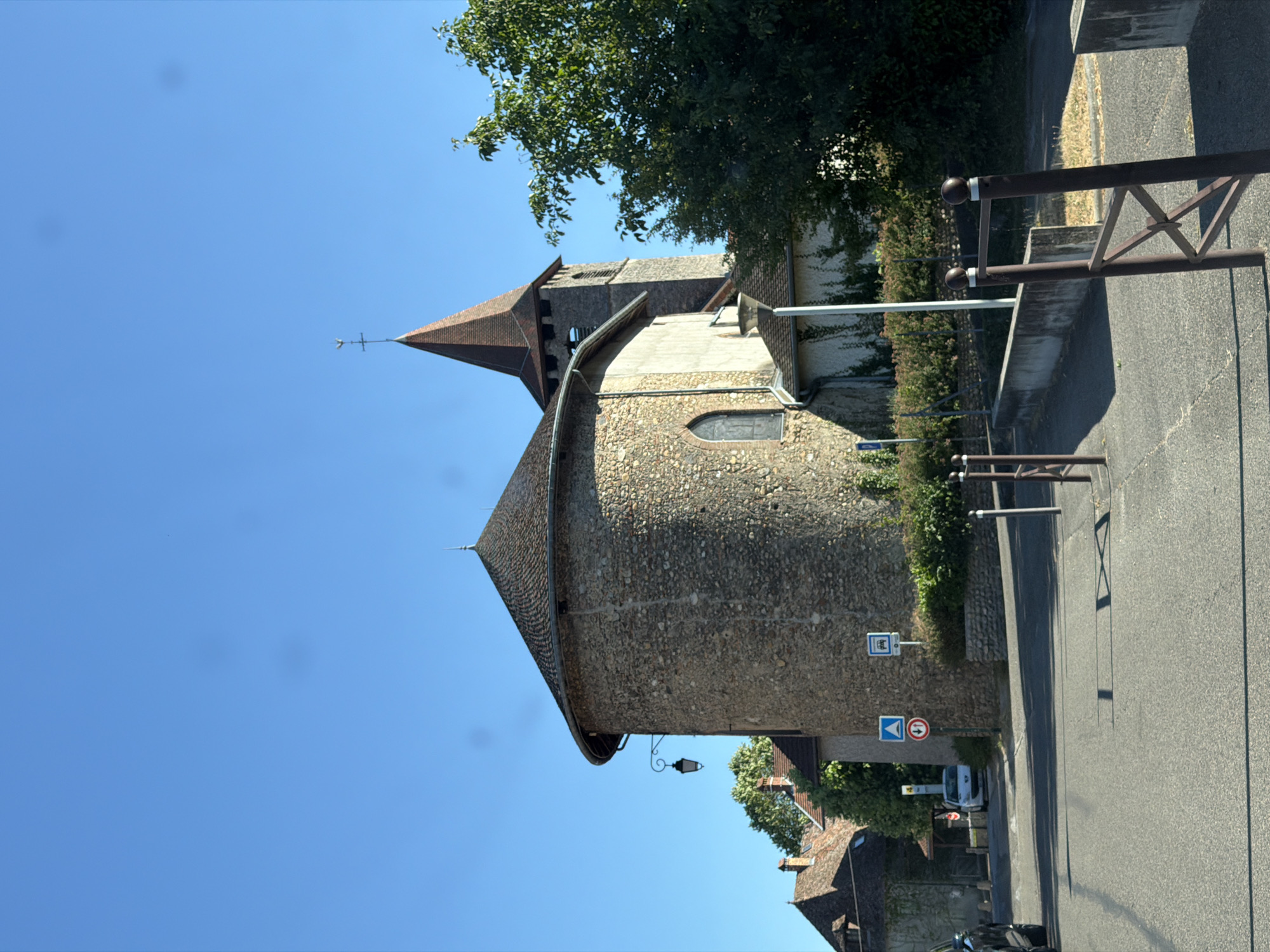 The image depicts a cylindrical stone building with a conical tiled roof, possibly a church or a historical tower, set against a clear blue sky. A pointed, brown-tiled roof with a weathervane crowns the structure. The stone walls are rough-hewn and appear aged. A small, arched window with a darkened pane is visible on the rounded facade. A decorative wrought-iron lantern hangs from a bracket below the roofline.
To the right of the building, a paved road runs parallel to a low stone wall, topped with a metal railing. Dense green foliage and trees are visible behind the wall. Several road signs are present: one blue and white sign with symbols, and another circular sign with a red border and a horizontal white bar, indicating a prohibition, possibly "no entry" or "no vehicles." Further down the road, a white van is parked.
The scene suggests a daytime setting, with bright sunlight casting shadows on the road. The weather appears clear and sunny. The architecture and signage hint at a location in a village or town, consistent with the provided context of Villages du Lac de Paladru, France. There are no people visible in the image, and the overall impression is one of a quiet, historical European setting. The orientation of the image is rotated 90 degrees clockwise.