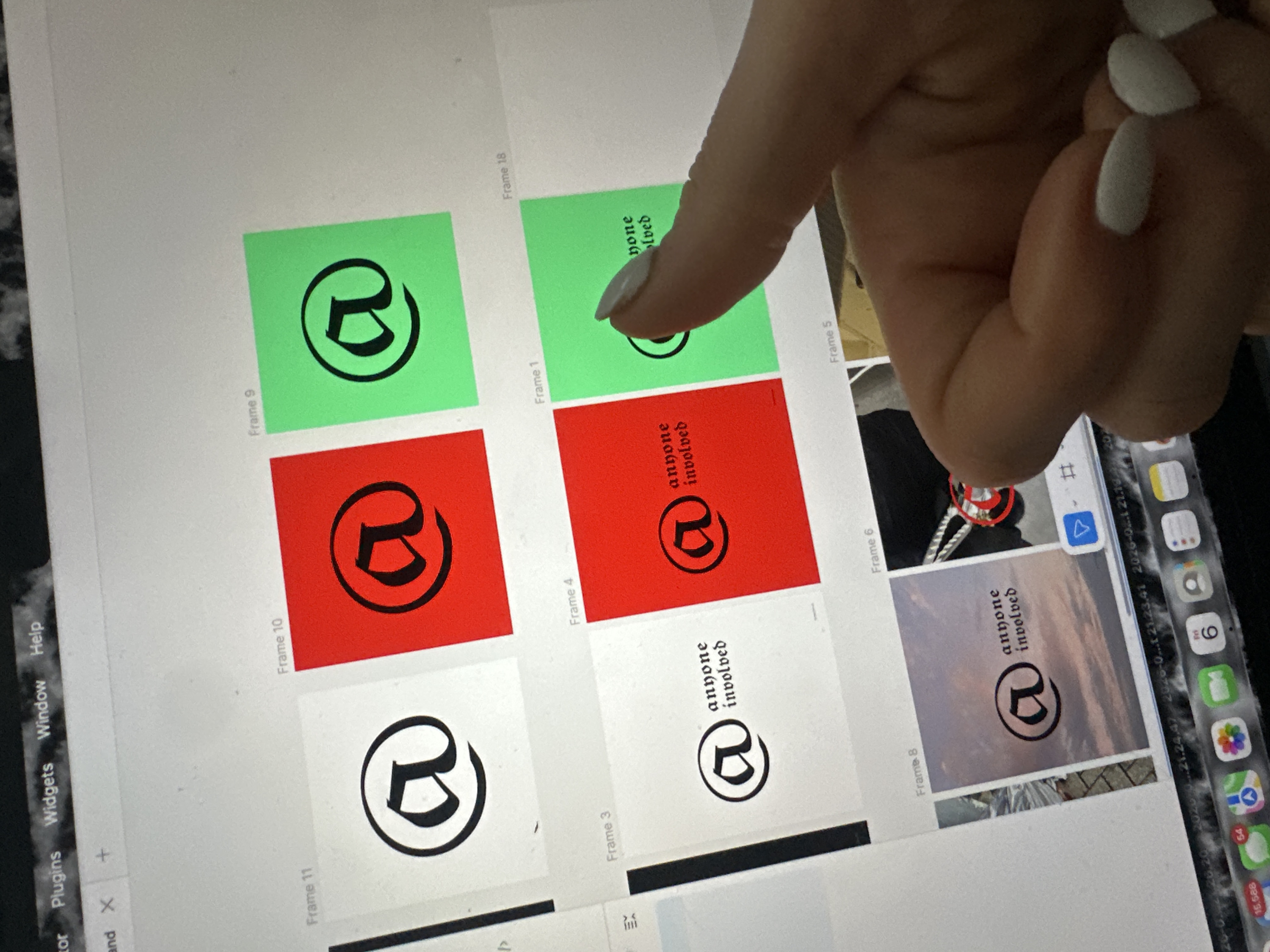 The scene, located in Tunbridge Wells, United Kingdom, captures a human hand interacting with a digital screen, likely a tablet or laptop, displaying a grid of graphical design elements within an application interface. The screen's top menu bar shows "Plugins," "Widgets," "Window," and "Help." The main display area presents multiple frames, each containing a black "@" symbol. Some frames additionally feature the text "anyone involved" in a gothic script. These frames utilize white, red, and green backgrounds. Specifically, Frame 11 is white with "@", Frame 10 is red with "@", and Frame 9 is green with "@". Below this, Frame 3 is white with "@" and "anyone involved," Frame 4 is red with "@" and "anyone involved," and Frame 1 is green with "@" and "anyone involved." A partial, empty white frame labeled "Frame 18" is visible to the far right. A human hand, with manicured white-tipped fingernails, is positioned on the lower right, with the index finger touching the green "Frame 1" area of the screen. The device's dock is partially visible at the bottom of the screen, showing application icons including Messages (with 54 notifications), Maps, Photos, and a calendar displaying "Fri 6." Faint timestamps or filenames such as "2020-0...21.24.47" are discernible above the dock. The surrounding environment appears dark and indistinct, reflecting light from the screen.