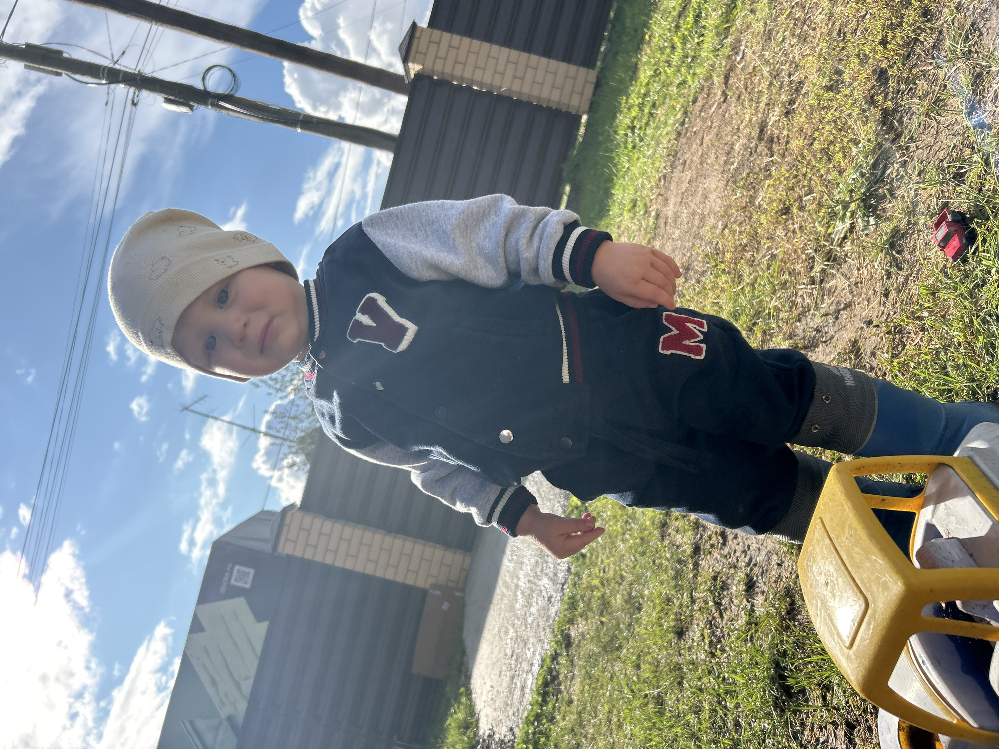 A young child, appearing to be around two years old, stands outdoors on a sunny day in Chaikovsky, Russia. The child is wearing a beige knit hat with a repeating cloud pattern, a dark blue and gray varsity jacket with a white "Y" emblem, and dark blue pants with a red "M" emblem. The child is also wearing blue rubber boots. In front of the child is a yellow plastic toy truck. The background shows a dark fence, a brick pillar, a building with a large graphic and a QR code, utility poles with wires, and a patch of grass. The ground is a mix of dirt and grass. The lighting suggests it is late afternoon.