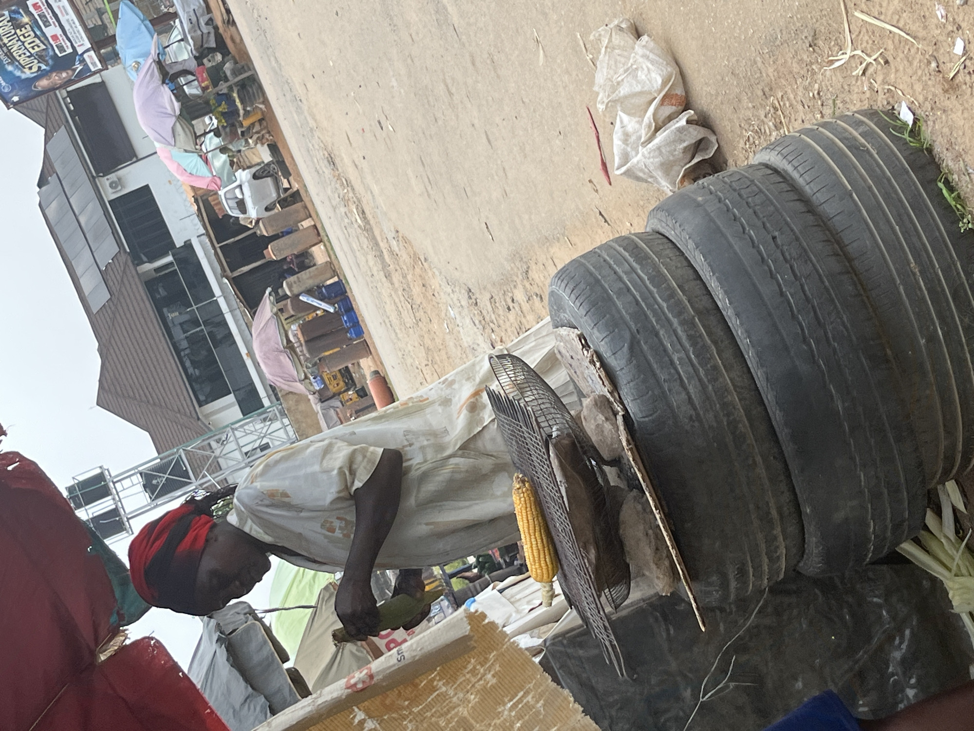 The image captures a street scene in Abuja, Nigeria, focusing on a woman roasting corn. She is standing under a red canopy, wearing a white garment and a red headscarf, and is in the process of tending to the corn on a makeshift grill. The grill appears to be constructed from a wire mesh, propped up by stones, and placed atop a stack of old tires. In the background, various market stalls are visible, with umbrellas providing shade. A building with solar panels on its roof can be seen further in the distance, suggesting a blend of modern and traditional infrastructure. The ground is dusty, and there are other vendors and customers in the periphery, though they are not the primary focus of the image. The overall atmosphere suggests a bustling open-air market typical of a Nigerian city.