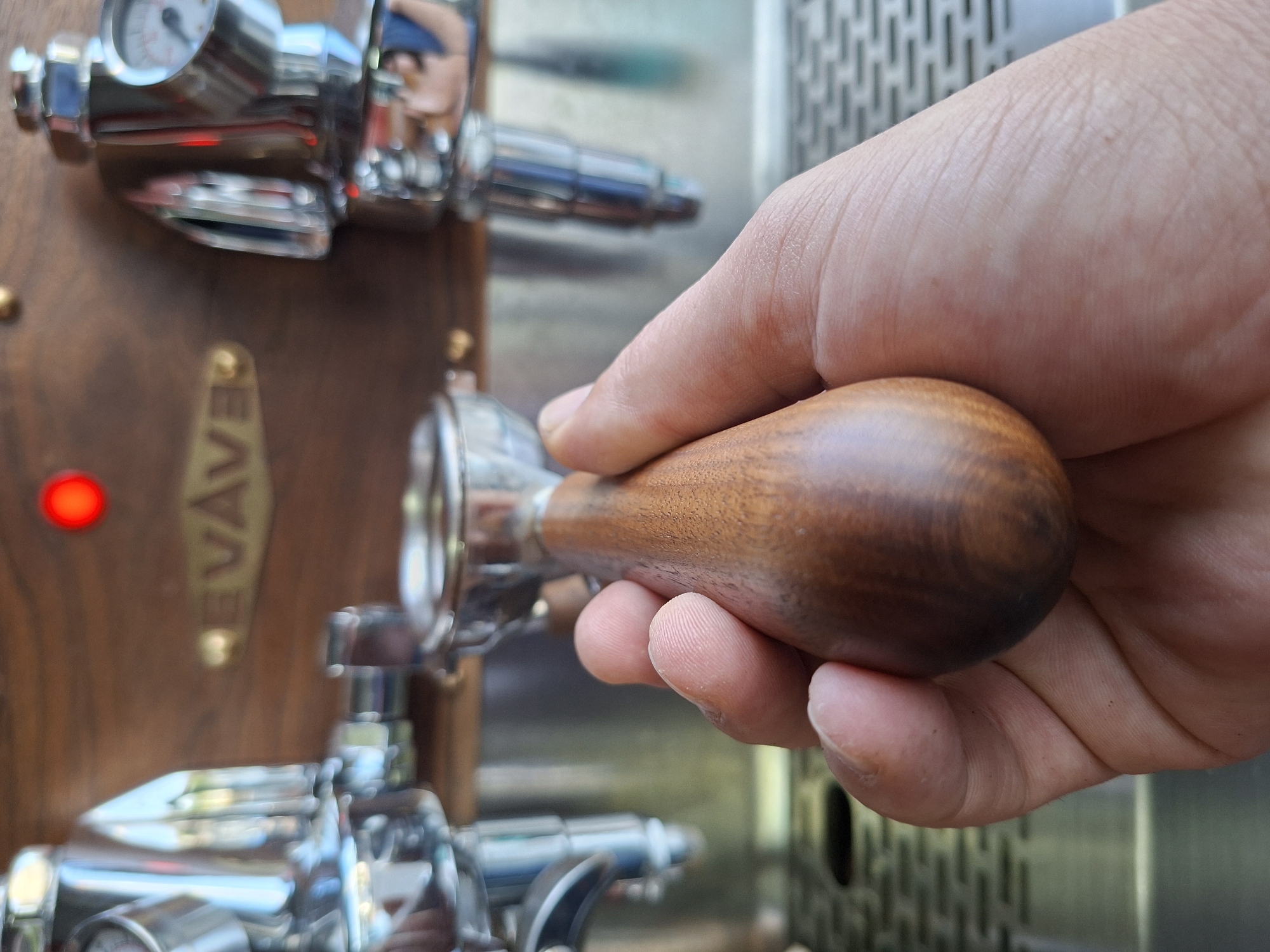 An individual's hand, exhibiting visible fingerprints and skin texture, is holding the wooden handle of an espresso machine's portafilter. The portafilter itself is made of polished metal. Behind the hand and portafilter, a portion of the espresso machine is visible, featuring a wooden panel with a circular red indicator light and a brass-colored emblem that reads "EVAWE". The machine also has chrome accents, gauges, and various metallic components. The background is blurred, suggesting a shallow depth of field, and includes metallic surfaces that reflect light and faint shapes of other objects. The image is taken from a close-up, low-angle perspective. The location is not specified in the image or its context.