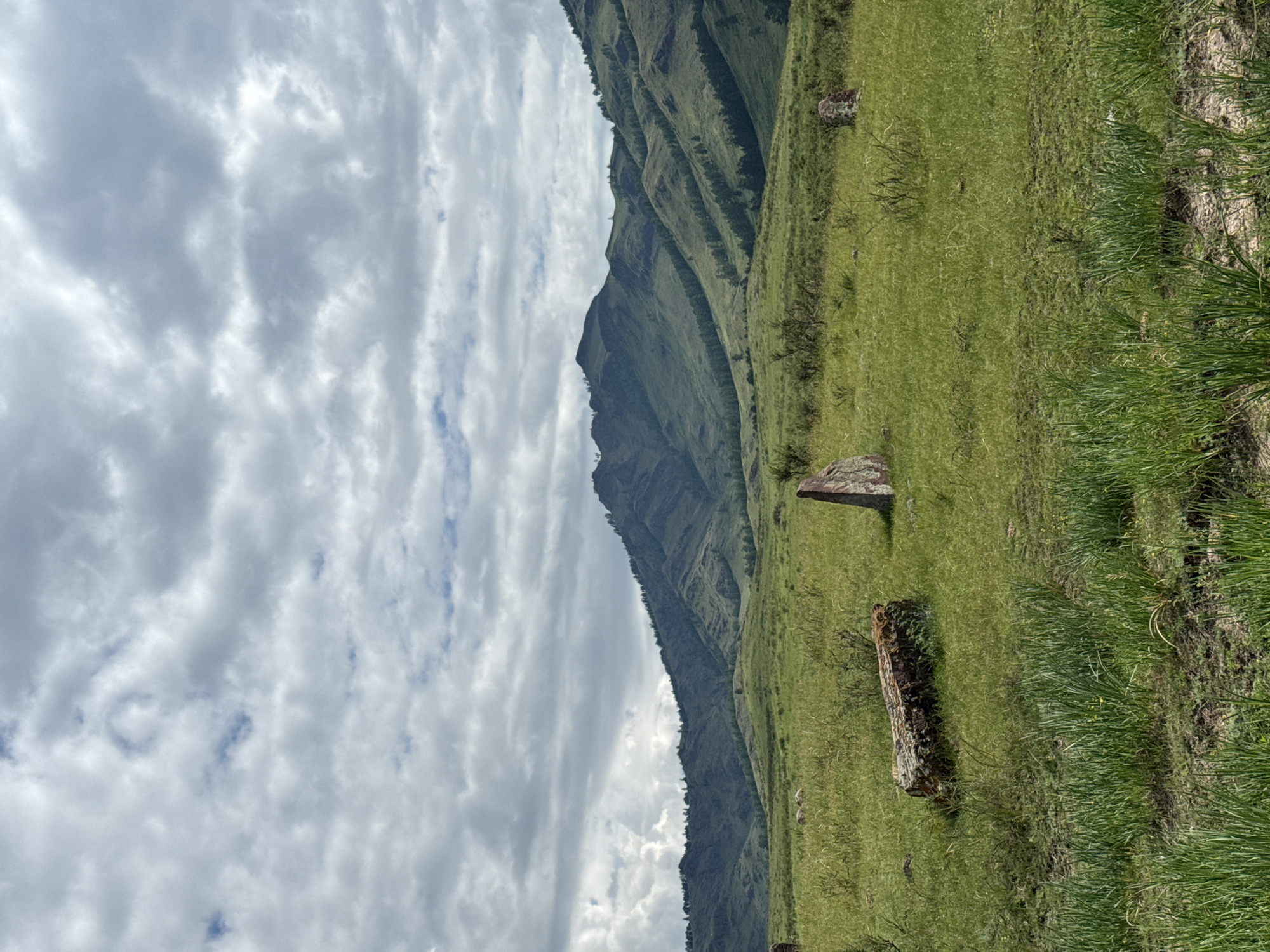 The image depicts a serene landscape featuring rolling green hills and mountains under a cloudy sky. In the foreground, several large, weathered stones are scattered across a grassy meadow. One prominent stone lies horizontally, covered in moss, while another stands vertically to its right. Further into the field, more stones are visible, including a tall, triangular stone. The background is dominated by the majestic, layered slopes of the mountains, dotted with sparse trees. The overall scene suggests a natural, possibly ancient, setting. The location is identified as Kazanovka, Russia. There are no people or animals visible in the frame.