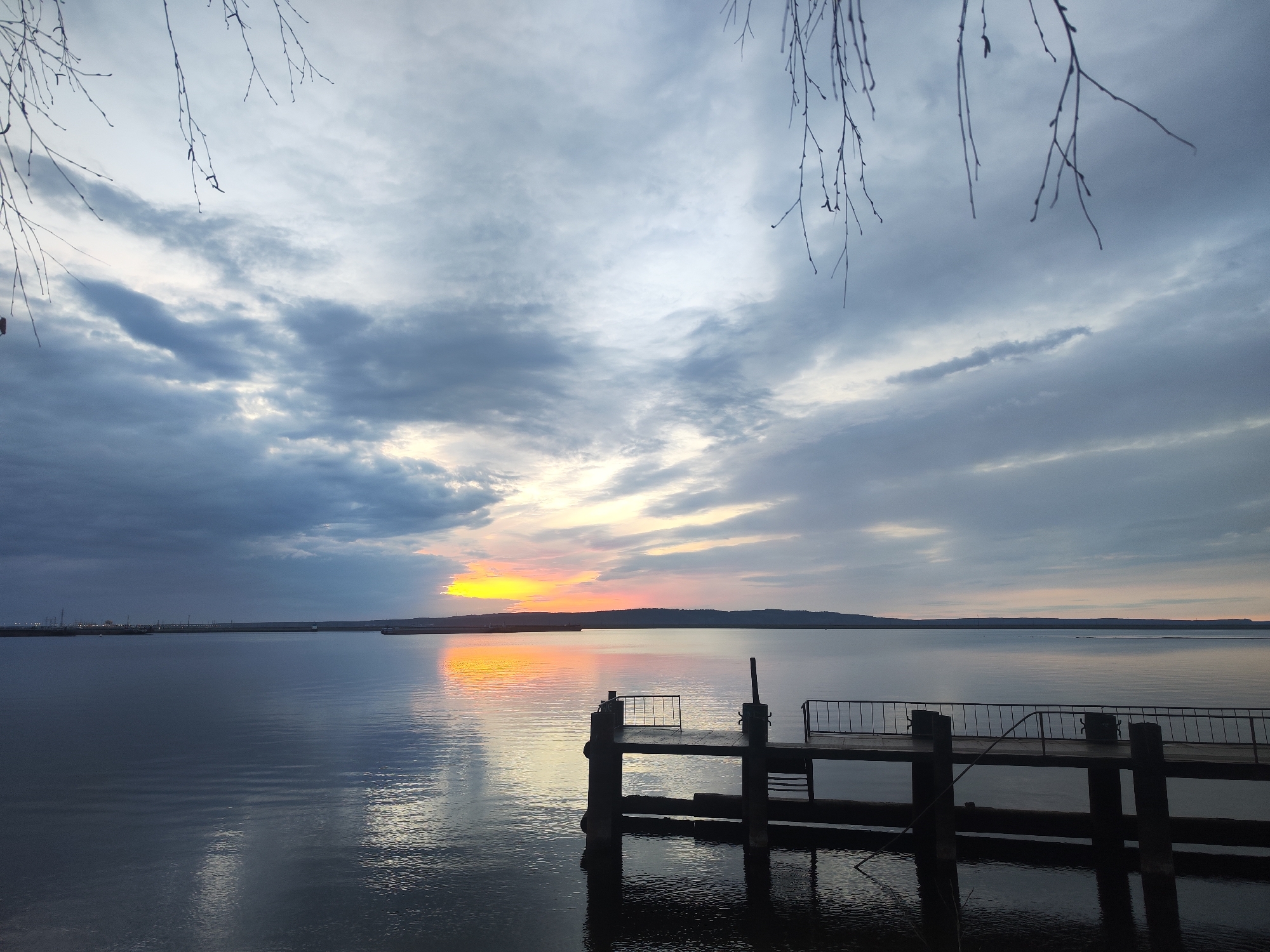 The image shows a sunset over the Kama Reservoir in Chaikovsky, Russia. A small wooden pier with railings juts out into the water from the foreground. The water is calm and reflects the colors of the sunset. In the background, a line of trees and possibly some industrial infrastructure is visible along the distant shoreline. The sky is cloudy with a bright orange and yellow glow near the horizon. Bare tree branches frame the top of the image, suggesting it's late autumn or early spring. There are no people visible in the image.