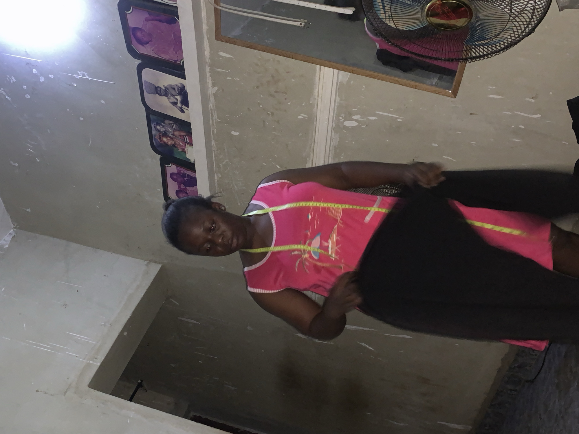 A woman in a pink sleeveless dress and a measuring tape around her neck is holding a piece of black fabric. She is standing in a room with unfinished walls, which are stained and have white splotches. On the wall behind her, there are framed photographs. To her right, a mirror reflects part of the room, and a fan is visible in the foreground. The scene appears to be in a residential area in Akwanga, Nigeria.