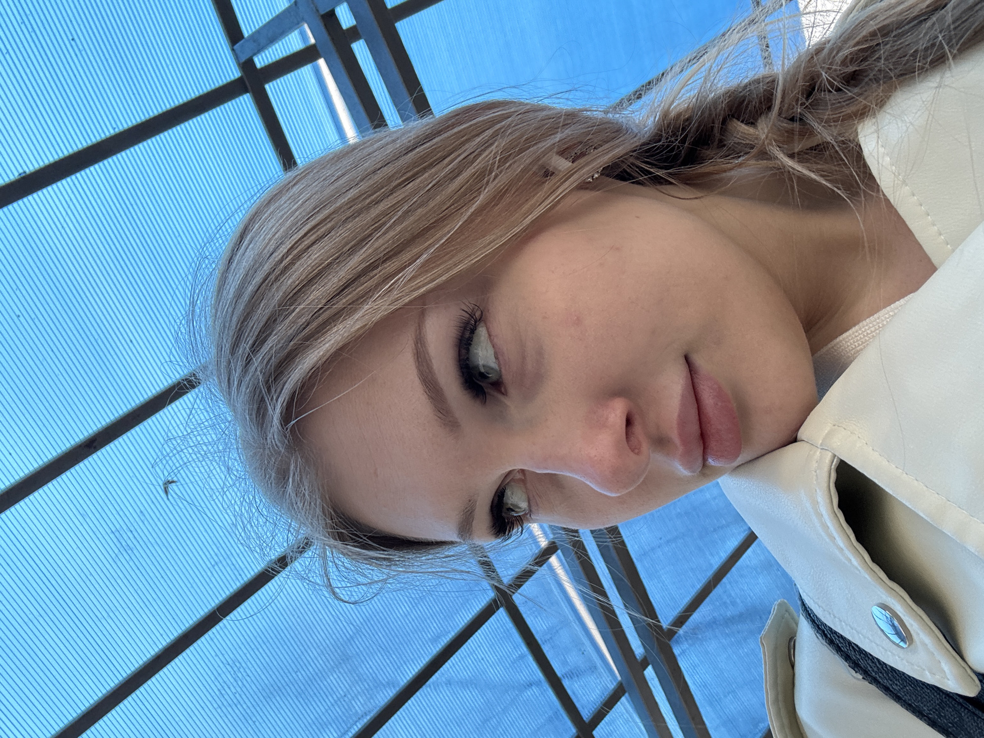 A young woman with blonde hair is lying down and looking up at the camera. She is wearing a white jacket. The background appears to be a blue, translucent roof structure with dark metal beams. Based on the image, it's impossible to definitively pinpoint the exact location within Perm, Russia. More contextual information would be needed for an accurate identification.
