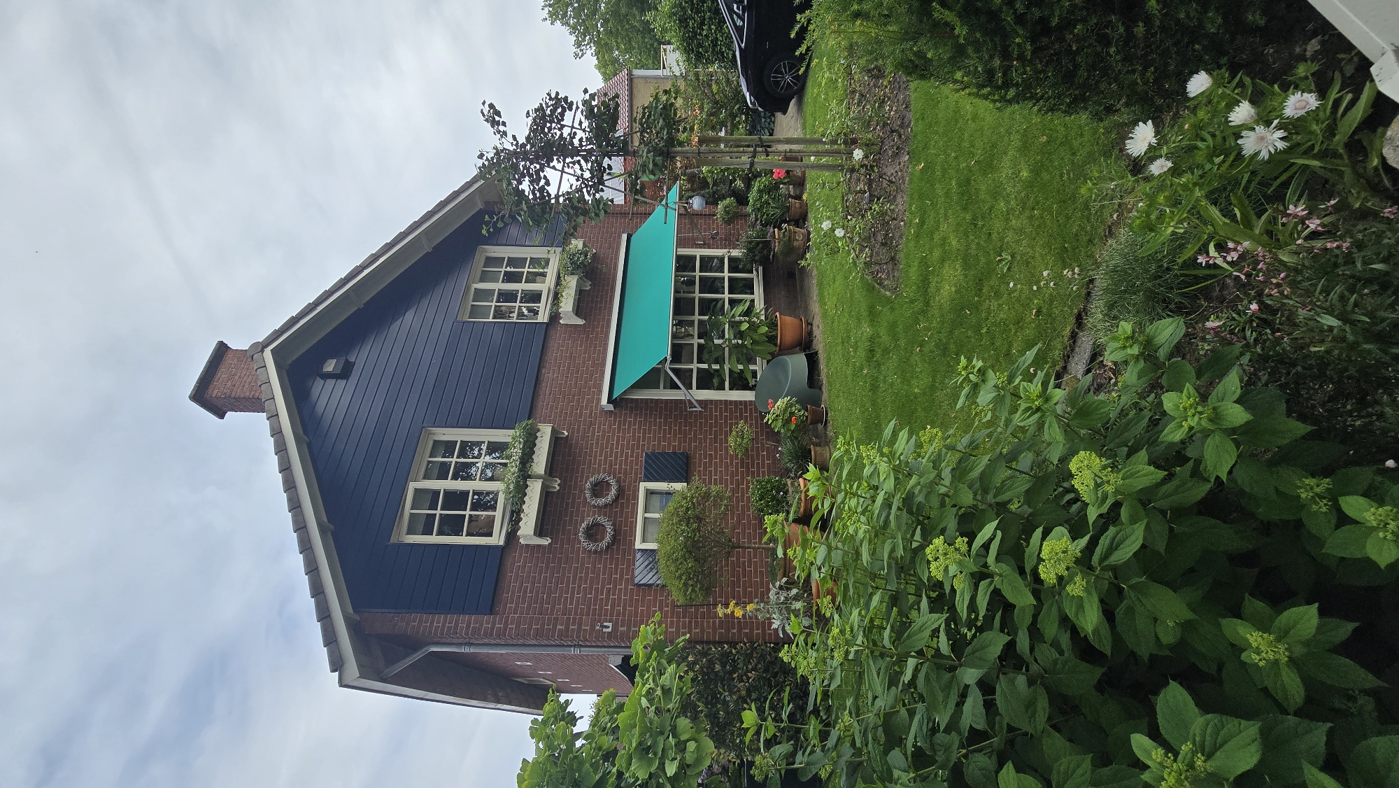 The image shows a brick house with dark blue siding on the upper level, located in Zeddam, Netherlands. A green awning extends over a large window on the first floor. The front yard is landscaped with a lush green lawn, various plants, and potted flowers. A dark-colored car is parked in the driveway to the right. The sky is overcast.
