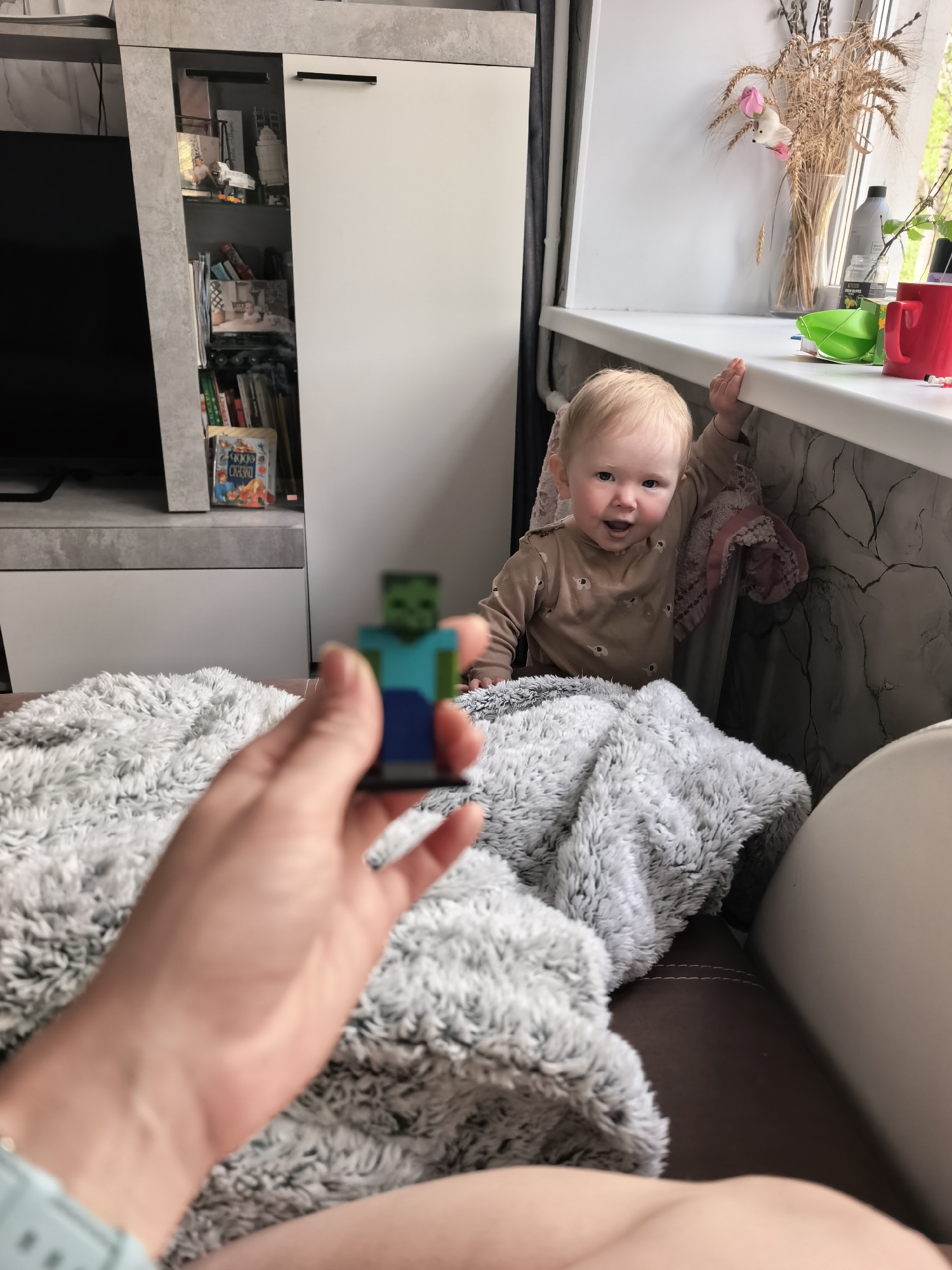 A person is holding a Minecraft Zombie figurine in the foreground, slightly out of focus. Behind them is a light gray fluffy blanket. A baby is standing by a window, holding onto the windowsill, and looking towards the camera with a smile. There are items such as flowers and a red mug on the windowsill. To the left, a white cabinet with a gray trim is visible. The wall near the window has a marbled pattern.
I do not have information to confirm the specific location of Sterlitamak, Russia.