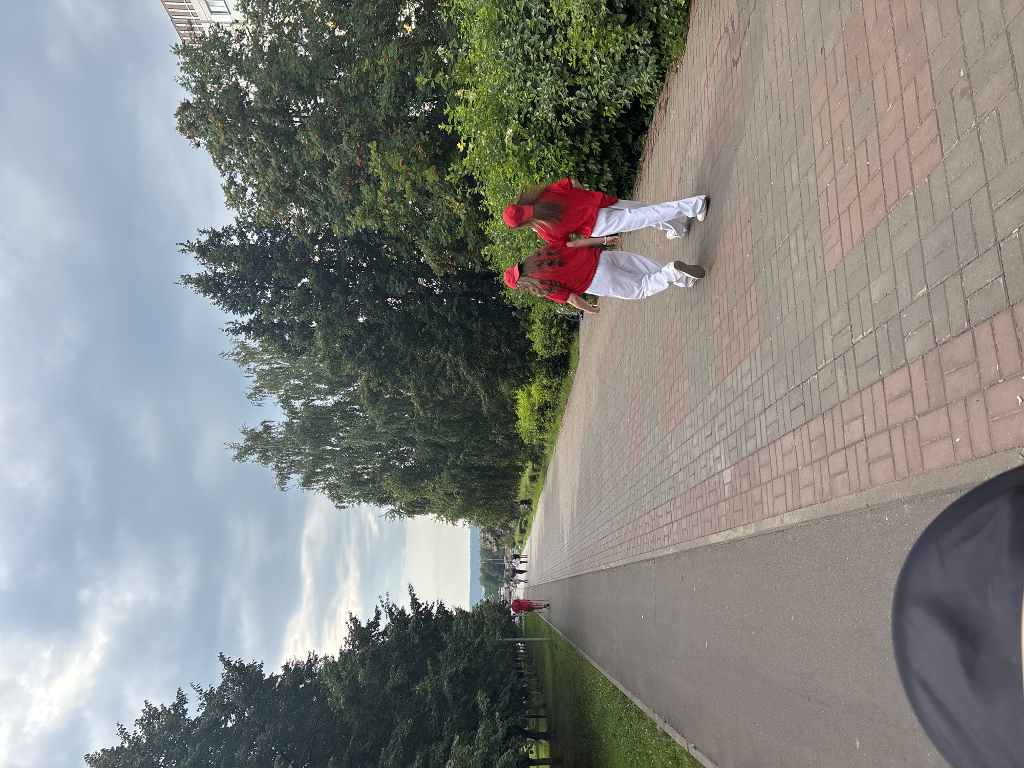 Two individuals, each wearing a red t-shirt and white pants with red baseball caps, are walking away from the camera on a paved walkway. The walkway is constructed with red and gray rectangular bricks laid in a herringbone pattern. To the left of the walkway is a paved path, likely for bicycles or scooters, bordered by a strip of green grass and then lined with mature trees. To the right of the walkway is a dense area of green bushes and trees, with a hint of a building visible in the upper right corner. The sky above is overcast with gray clouds. In the distance, other people can be seen walking on the walkway. The scene appears to be a park or a pedestrian zone within a city, likely in Naberezhnye Chelny, Russia, given the context.