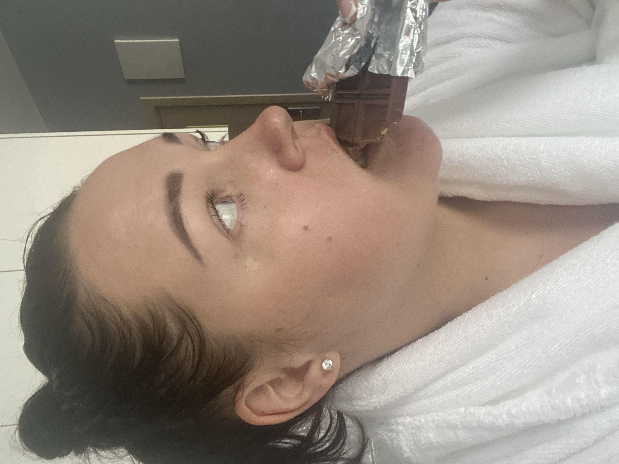 A woman in a white robe is eating a large piece of chocolate that is partially wrapped in silver foil. She is indoors. It is not possible to determine if this is Sochi, Russia.
