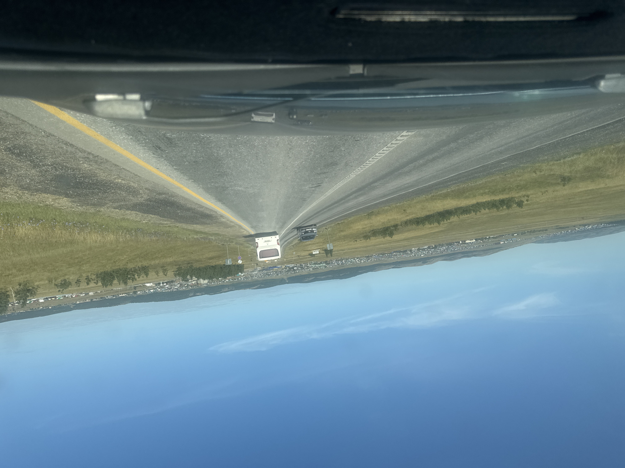 The image captures a view from a vehicle driving on a paved road in a rural or suburban area. The road has two lanes in the direction of travel, with a broken white line marking the lanes and a solid yellow line on the right edge. In the distance, a white bus and a dark-colored car are visible on the road. The surrounding landscape consists of rolling hills covered in dry grass and sparse vegetation, with a town or village spread out along the base of the hills. The sky is clear and blue, with a few wispy clouds. The foreground shows the dashboard of the vehicle, including the windshield and parts of the interior. The scene appears to be in Baymak, Russia, based on the provided context.