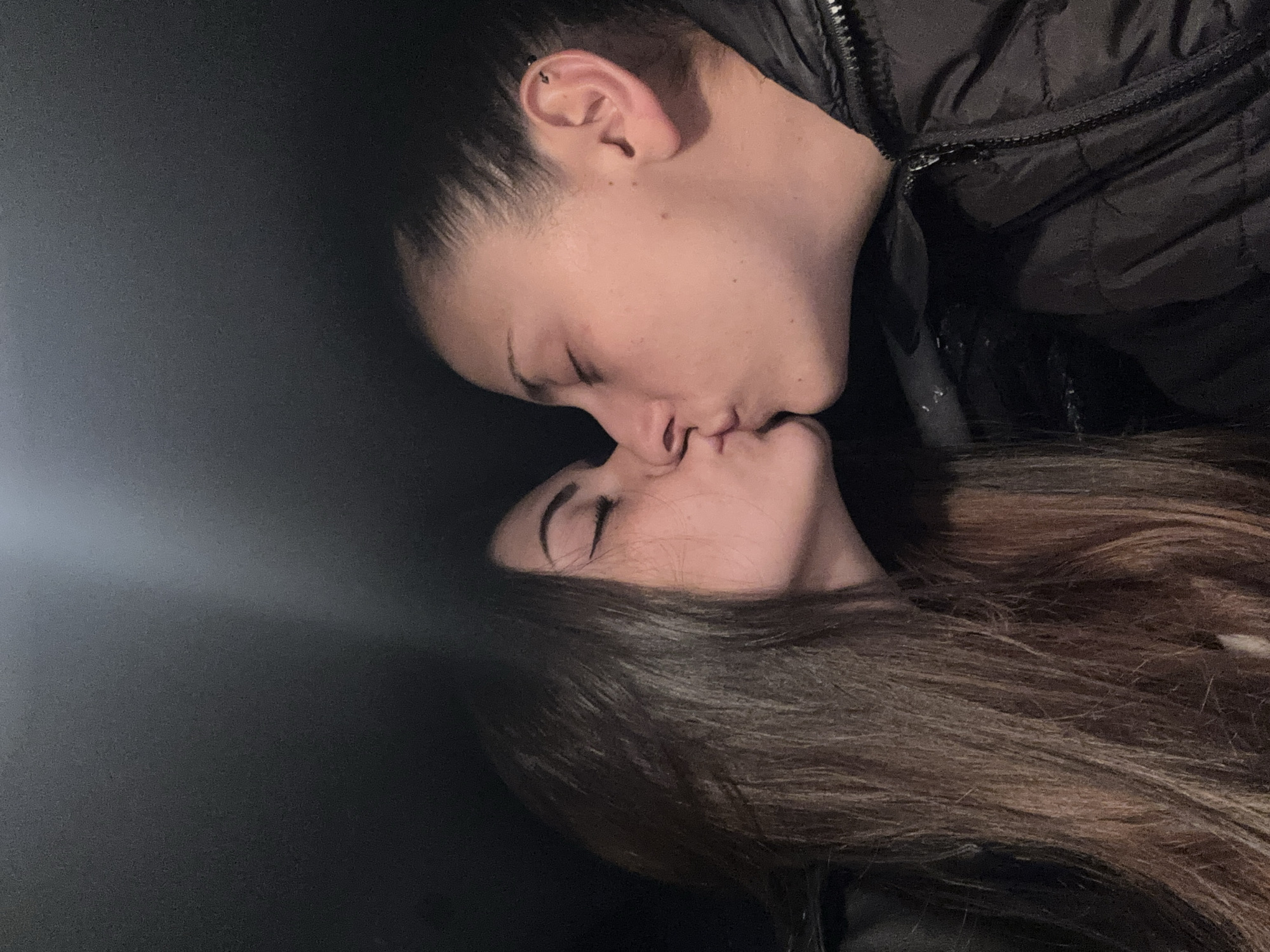 Two people are kissing. One person has short dark hair, a piercing in their ear, and is wearing a black jacket. The other person has long brown hair and is wearing a black jacket. The background is dark. There is no evidence to identify the location as Sofia, Bulgaria.