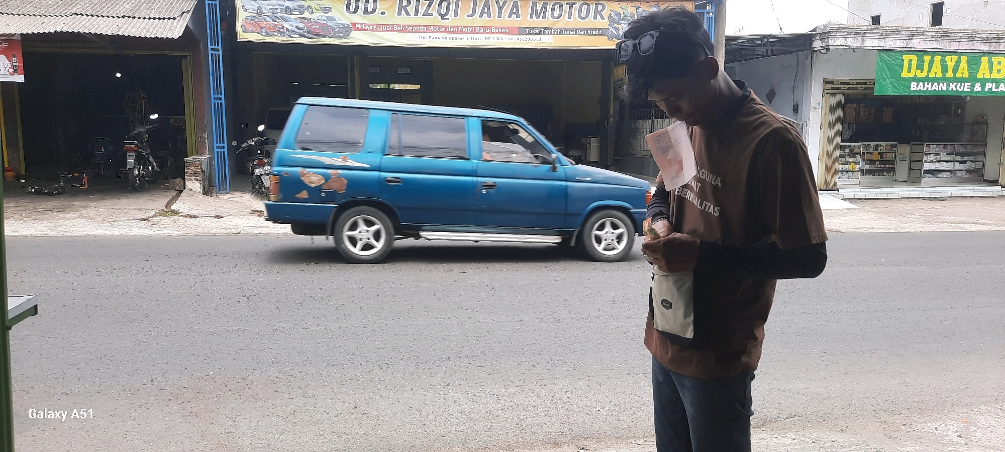 The image captures a street scene in Selopuro, Indonesia, featuring a blue Isuzu Panther van driving on the right side of a paved road. The van exhibits visible rust spots, particularly on its rear side. In the foreground, a young man, wearing a brown t-shirt over a dark long-sleeved shirt and blue jeans, is standing on the sidewalk, looking down and holding a small paper and what appears to be a wallet or coin purse. He has dark hair and sunglasses perched on his head.
Behind the van, a building with a sign that reads "UD. RIZQI JAYA MOTOR" is visible, indicating a motorcycle and car sales and service shop. The shop has an open garage entrance where several motorcycles are parked. To the right of the motor shop, another building houses a store with a green banner that says "DJAYA ABADI BAHAN KUE & PLASTIK," suggesting it sells baking ingredients and plastic goods. This store features glass display shelves stocked with various products. The environment appears to be a commercial area with multiple businesses lining the street. The weather is overcast.