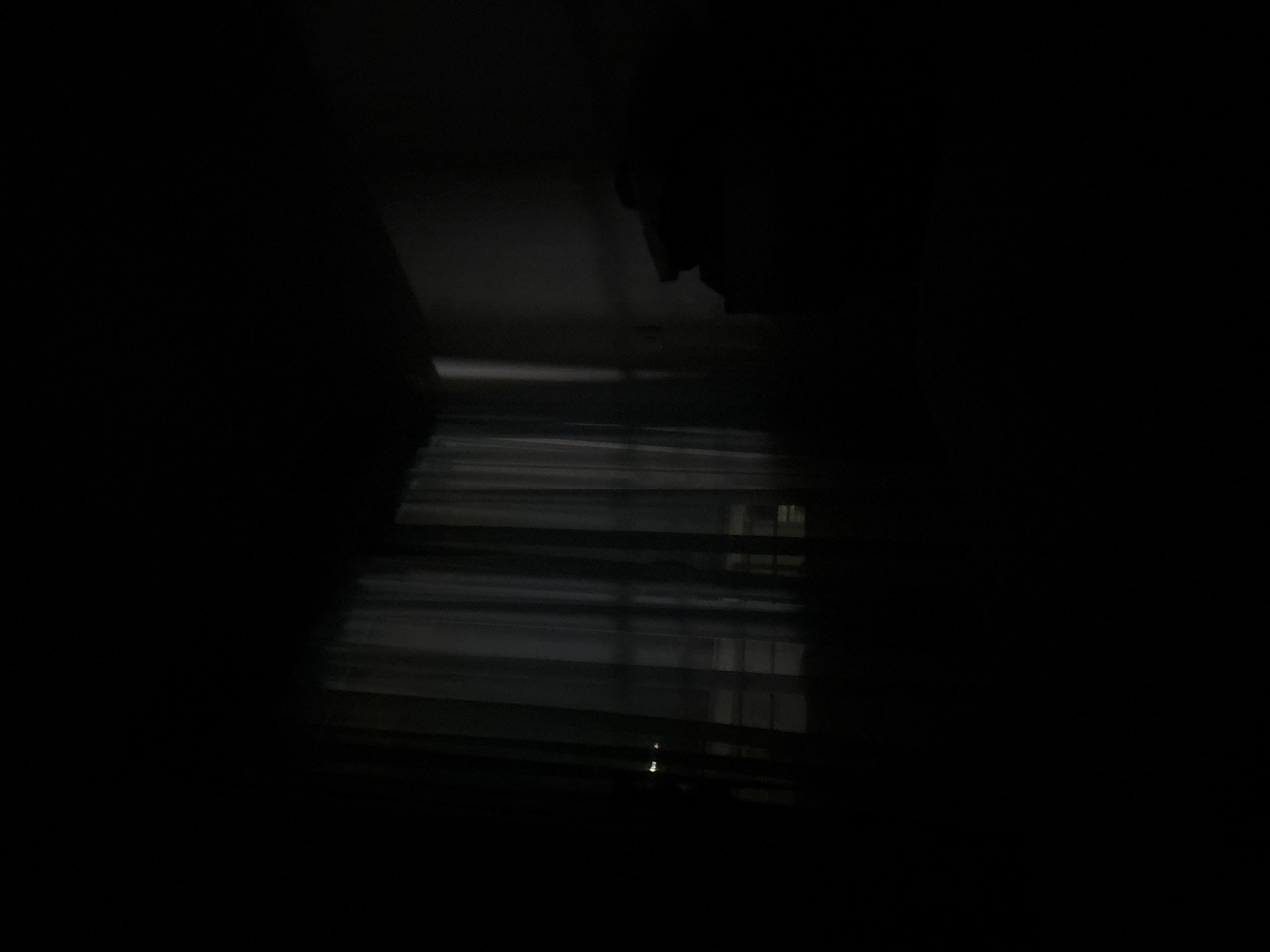 The image shows a dark room with light filtering through closed blinds. Outside the blinds, there appears to be another building visible. The details are difficult to discern due to the darkness. The location is not identifiable as Budva, Montenegro, from the visual data provided.