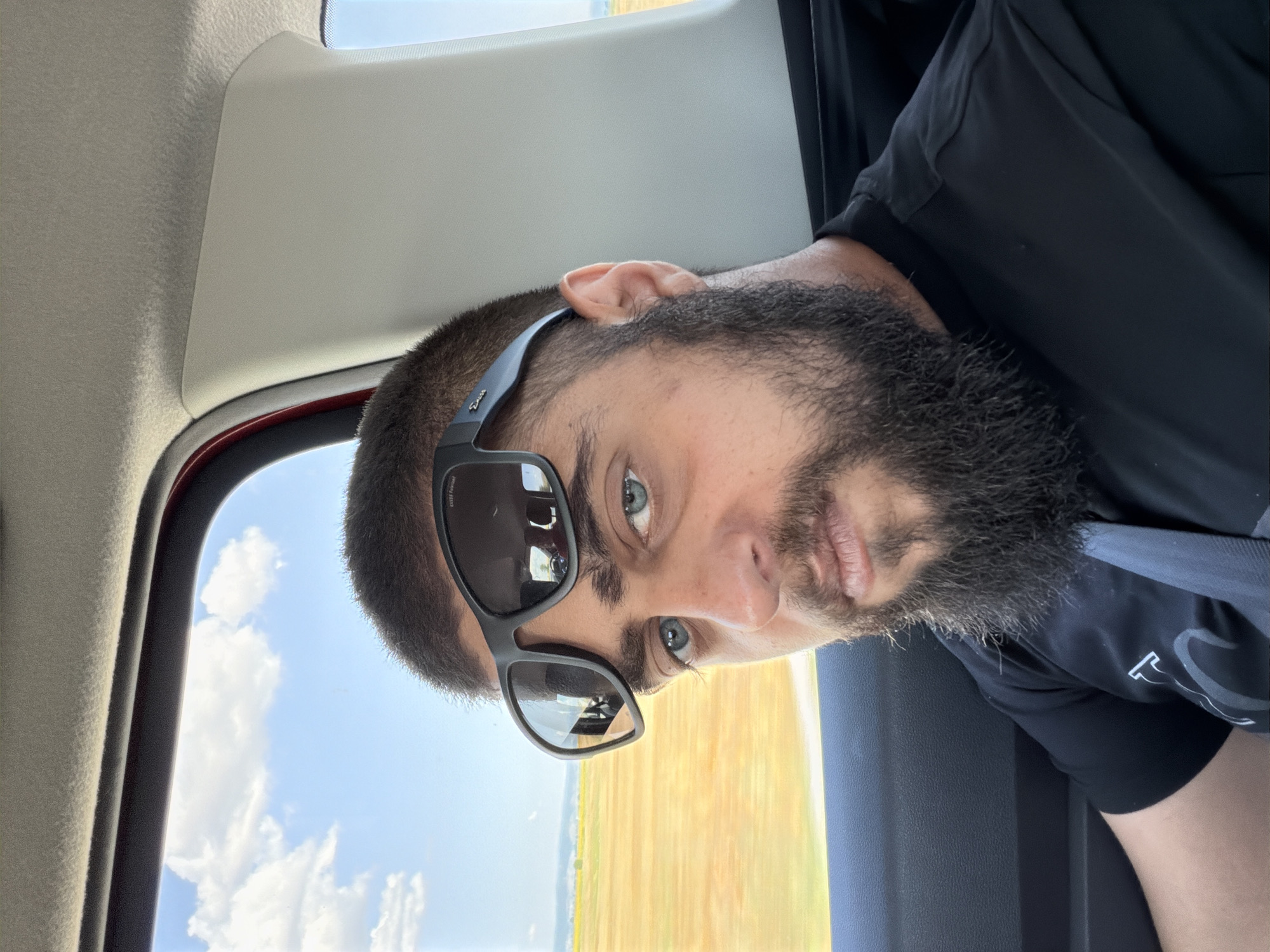 A close-up selfie shows a man with a beard and blue eyes wearing sunglasses. He is sitting in a car, with a window to his left showing a blurred landscape of yellow fields and a blue sky with clouds. The interior of the car is visible, including the headliner and a black seatbelt. The man is wearing a black t-shirt. The image appears to have been taken while the car was in motion.