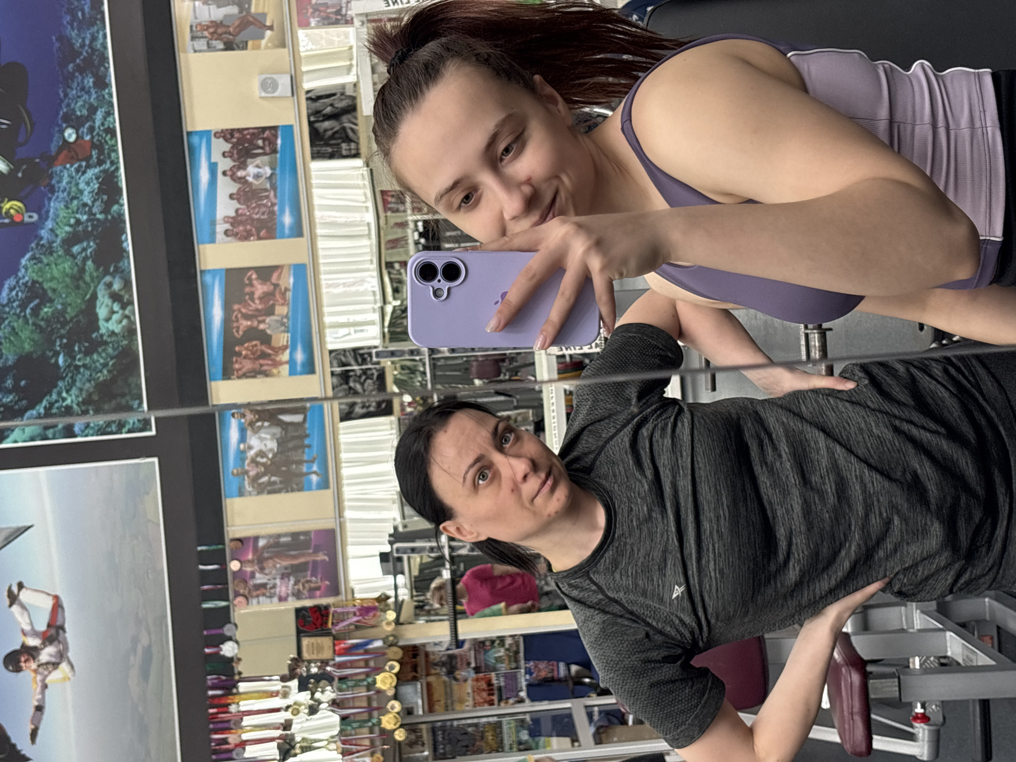 A young woman with dark hair pulled back in a ponytail takes a selfie in front of a mirror. She holds a lavender-colored smartphone. She is wearing a purple sports bra and a gray tank top. The reflection in the mirror shows another woman with darker hair wearing a gray athletic shirt. The background includes posters, shelves, and trophies. The location is Chaikovsky, Russia.