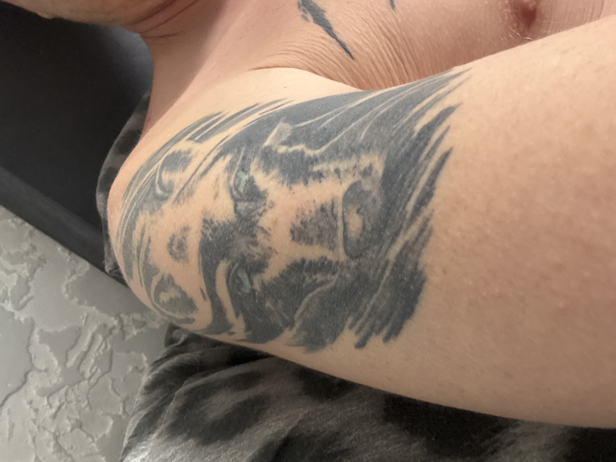 A close-up, head-and-shoulders shot focuses on a tattoo of a wolf's face on a person's upper arm and shoulder. The tattoo is rendered in black and grey, with some shading and detail in the wolf's fur and expression. The person is wearing a dark, possibly grey and black striped shirt on their left arm, which is partially visible. The background includes a textured, light-colored surface, possibly a wall, and a dark upholstered surface, perhaps a chair or couch. The image appears to be taken indoors, with natural or artificial lighting. There is no indication of the location being in Tolyatti, Russia.