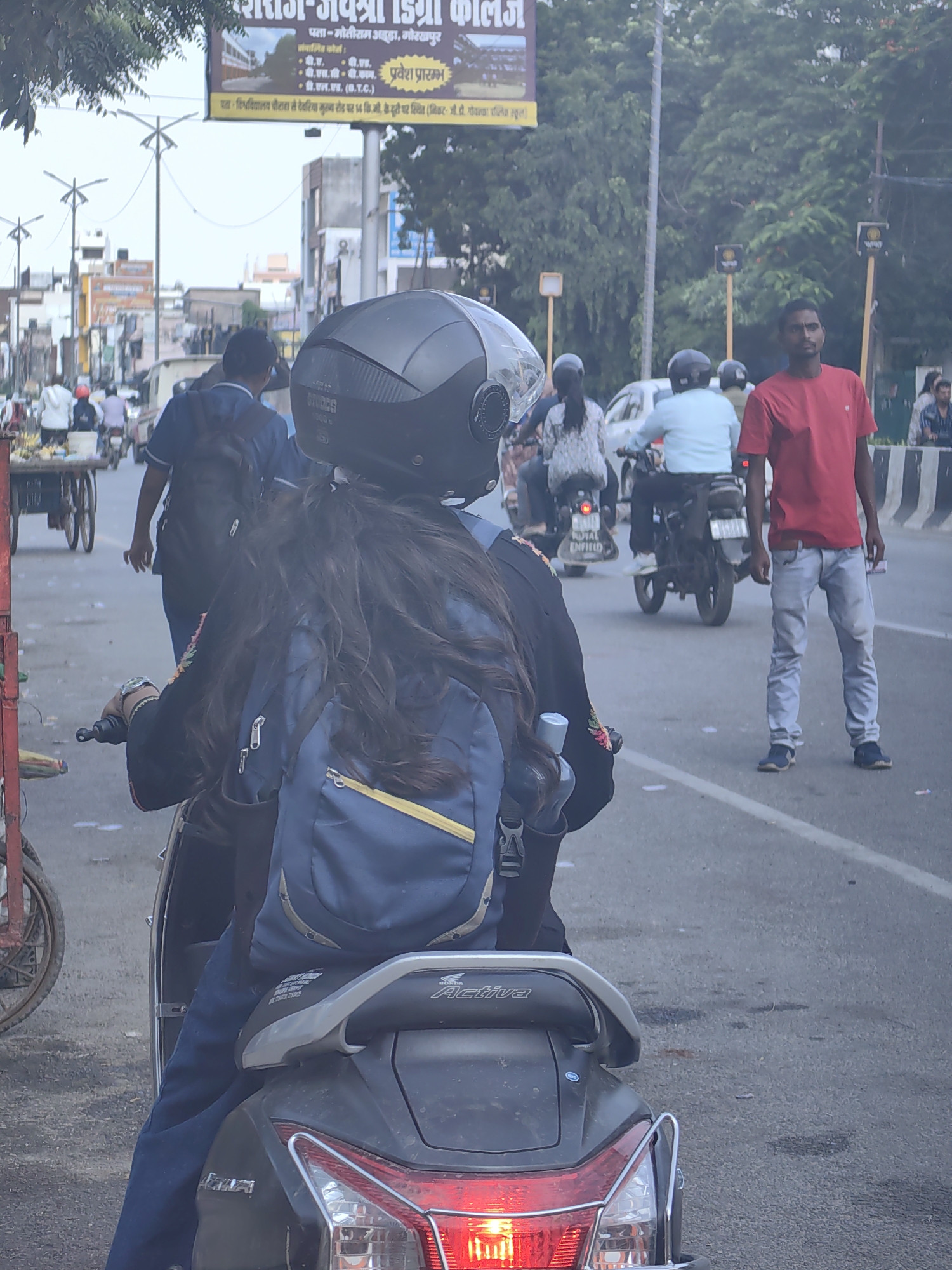 The image depicts a street scene in Gorakhpur, India, likely during the daytime, with a moderately busy road. In the foreground, a person is seated on a scooter, viewed from behind. This individual wears a dark jacket with embroidered floral patterns and a blue backpack is visible. The scooter's rear light is illuminated.
Further down the road, several other individuals and vehicles are present. A man in a red t-shirt and light-colored jeans stands on the right side of the road, facing towards the viewer. To the left and center, people are riding motorcycles. One motorcycle has two riders, one wearing a light blue shirt. Another motorcycle has a rider in a dark blue shirt and a backpack. In the background, there are buildings and trees lining the street, and a billboard with Hindi text is visible at the top. The road surface appears worn. The overall atmosphere suggests a typical urban environment in India.