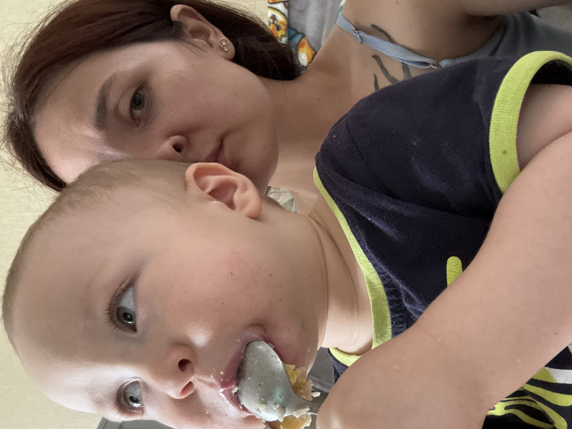 The image shows a close-up of a baby being fed by a woman. The baby is in the foreground, looking to the left, with a spoon in its mouth. The baby has some food on its face and is wearing a dark blue shirt with a neon green trim. Behind the baby, the woman is partially visible, looking towards the camera. She has dark hair and is wearing a light blue strap. The background appears to be an indoor setting, with a blurry bed or couch visible. There are no visible landmarks or indicators that would place this image specifically in Naberezhnye Chelny, Russia.
