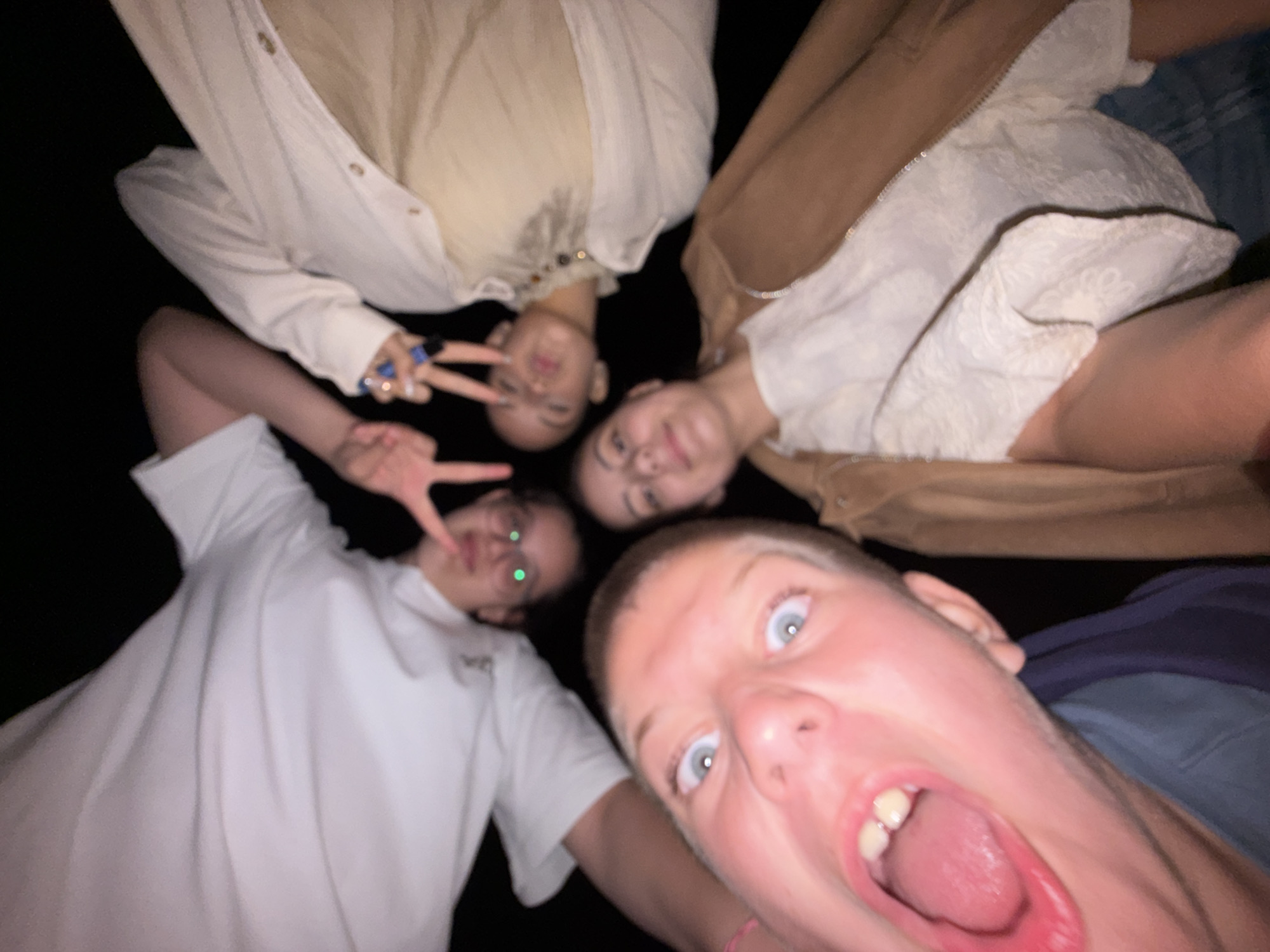 Four individuals are captured in a close-up, wide-angle group photo, arranged in a circular formation around the camera against a uniformly dark background, indicative of a low-light environment in the city of Tsarevo, Bulgaria.
The individual at the top, a male, is wearing a white long-sleeved shirt and glasses, displaying a peace sign with his left hand. His eyes show green reflections. To his right, a female is depicted wearing a light cream-colored long-sleeved shirt or jacket over a tan top. She is making a peace sign with her right hand, revealing rings on her fingers and blue nail polish. The individual positioned prominently at the bottom, a male, has light-colored short hair and wide blue eyes; his mouth is open wide, revealing his teeth and tongue, and he is wearing a dark blue garment. To the left, a female is partially visible, wearing a light-colored, possibly textured or embroidered top under a brown suede-like jacket. All individuals are looking towards the camera.