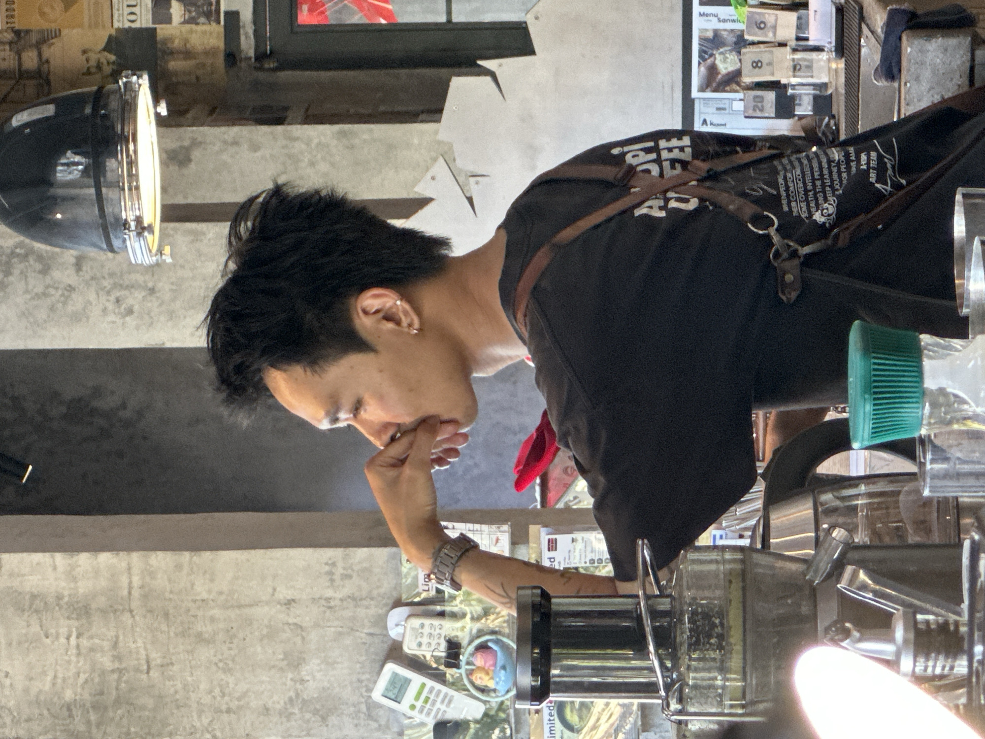The image shows a young man with black hair, wearing a black t-shirt and an apron, working behind a counter in what appears to be a cafe. He is holding his hand to his mouth, looking to his right. In front of him are various items of equipment, including a juicer, a kettle, and some containers. The background shows a concrete wall with some posters and a light fixture. The cafe is located in Da Nang, Vietnam.