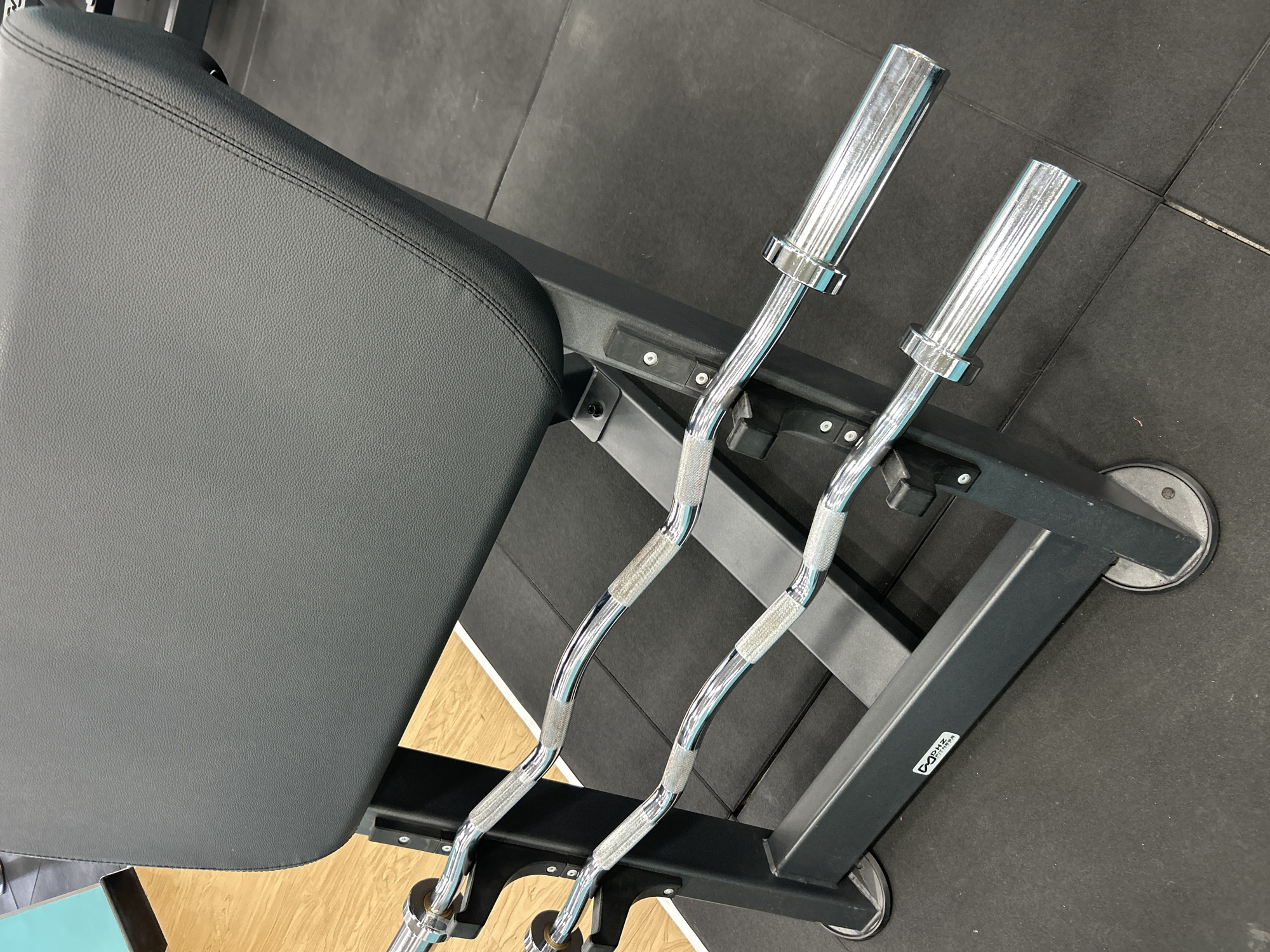 The image provides a high-angle view of exercise equipment within a gym or fitness facility, located in Kuta Selatan, Indonesia. The primary subjects are a dark gray or black padded workout bench and two shiny chrome EZ curl bars.
The padded bench, featuring a textured, leather-like surface with visible stitching, takes up a significant portion of the left side of the frame. It connects to a sturdy, black metal frame that also serves as a storage rack for the barbells. The frame includes wheels at its base, suggesting the equipment is movable.
Two gleaming chrome EZ curl bars rest securely on the black metal frame. These bars are characterized by their wavy shape and textured, knurled sections designed for improved grip during weightlifting. The smooth sleeves at the ends of the bars are where weight plates would be loaded.
The flooring in the scene consists mainly of dark gray or black rubber gym mats, which extend beneath and around the equipment. A section of lighter wooden flooring is also visible, hinting at different zones within the facility.
No people are visible, and no activity is actively taking place; however, the setup clearly indicates a space for weight training, particularly arm exercises. The lighting is bright and even, consistent with an indoor gym environment. A white rectangular sticker on the black metal frame explicitly displays the text "DAPHNE FITNESS," identifying the brand or establishment. The overall scene suggests a well-maintained and organized fitness area.