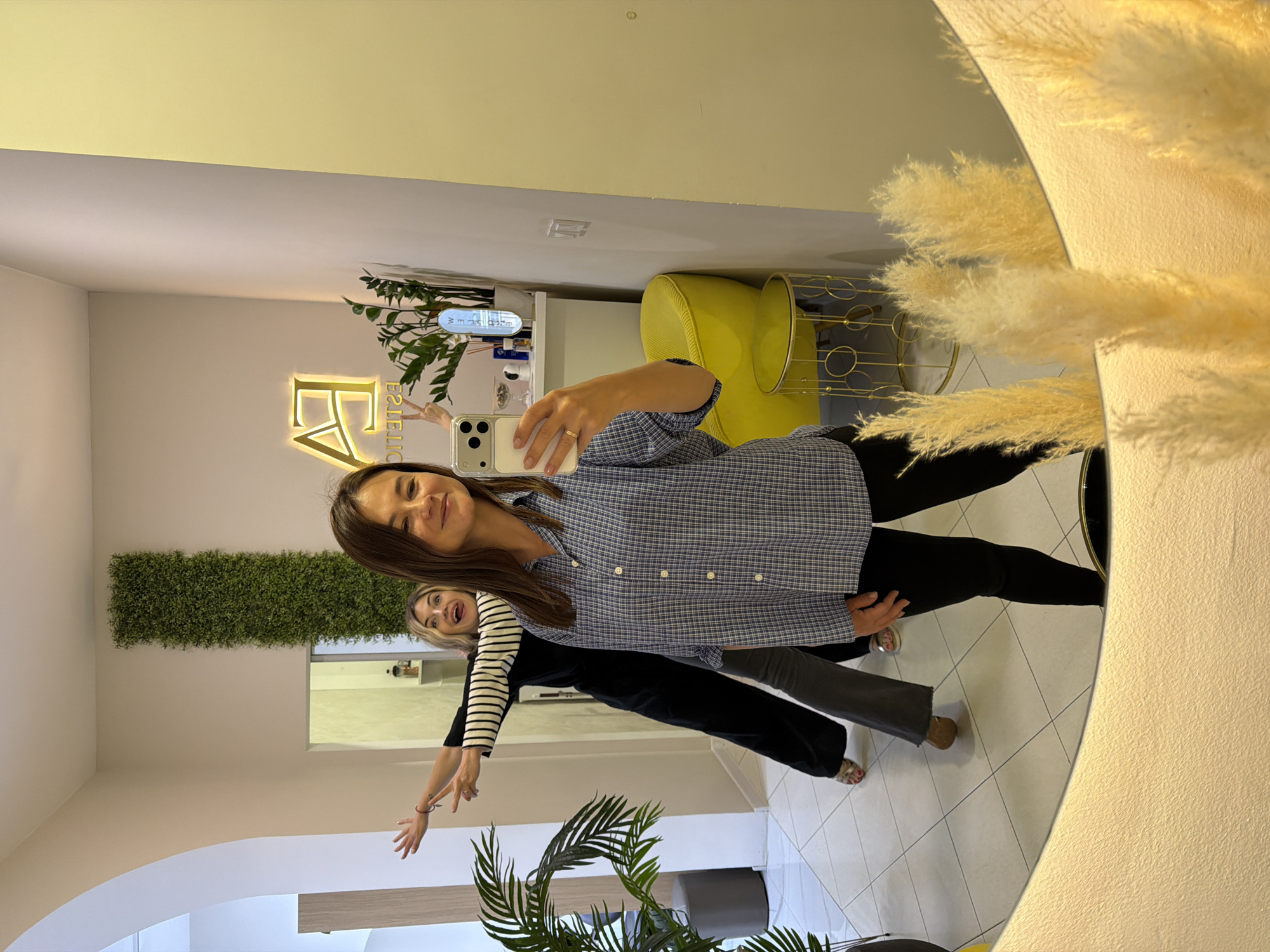 A medium indoor shot, in a commercial space, showing the reflection of two women taking a selfie in Budva, Montenegro. The primary woman, in the foreground, is light-skinned and has brown hair and is wearing a blue and white plaid button-down shirt and black pants. She holds a cell phone in her right hand. A second woman, visible behind her, has blonde hair and wears a black and white striped long-sleeve shirt and dark grey pants. She has both arms extended out towards the camera. A rectangular patch of artificial greenery hangs high on the wall behind them. An illuminated logo is on a wall, reading “FA Estetic”. Further behind them is a yellow chair and a white counter top with bottles. The scene is reflected in a large, round mirror with a fluffy decoration on the right. The floor is light-colored tile. A potted palm is visible below the mirror.